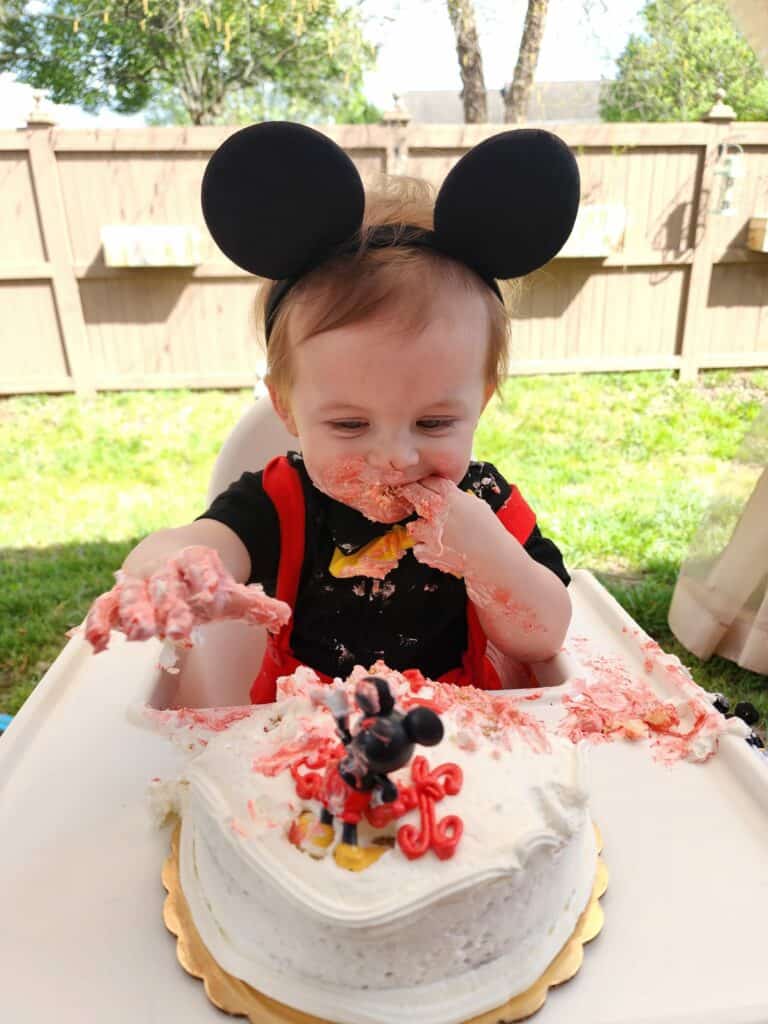 image of a toddler sitting in a high chair with a mess of a birthday cake before him. he is dressed like mickey mouse complete with ears. He is smiling and has frosting on his face. a 1st birthday party