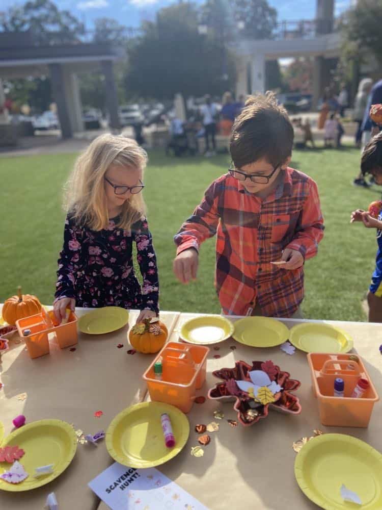 Two children engaging in a craft activity at an outdoor birthday party in Raleigh, NC, with a table set with yellow plates, orange containers, and autumn decorations under the sunny skies