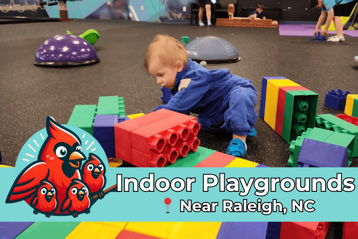 A young child in a blue jumpsuit plays with oversized colorful building blocks at an indoor playground near Raleigh, NC, with other children and activities visible in the background.
