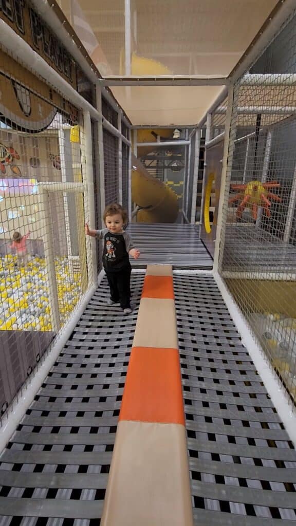 A curious toddler walks through a narrow bridge inside a mesh-enclosed play structure, with a soft padded floor for safety, at an indoor playground