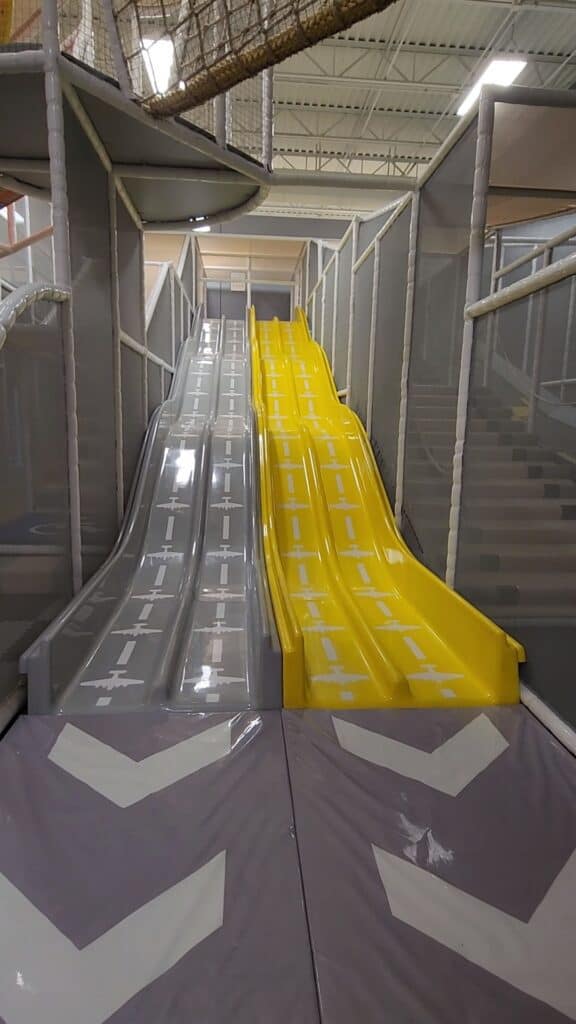 Dual plastic slides in gray and yellow await eager children for a fun ride down in a spacious, brightly lit indoor play area.