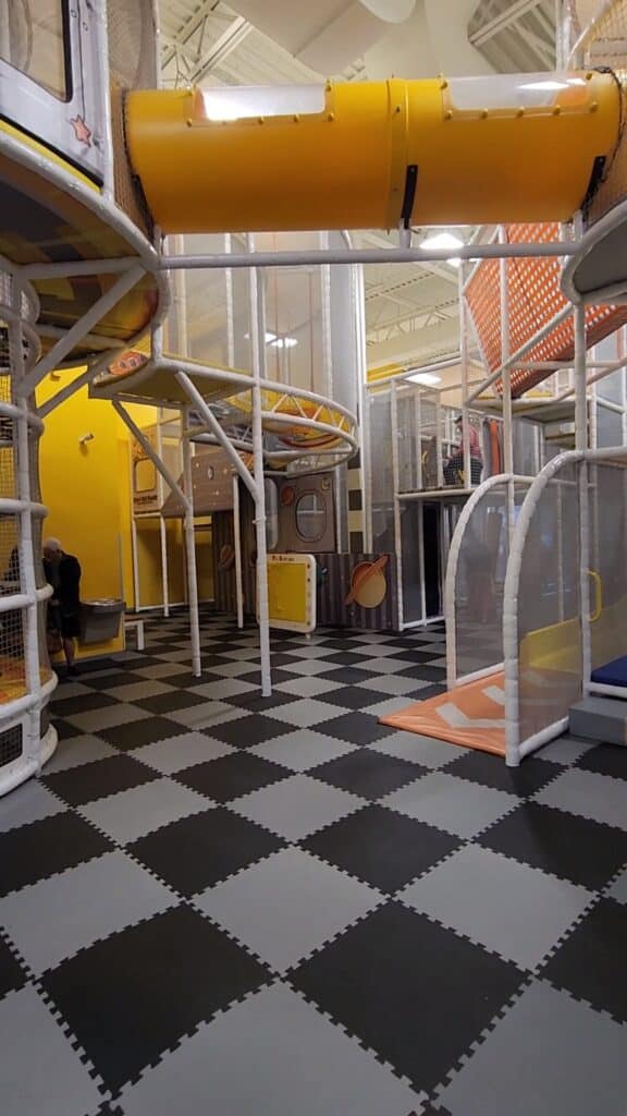 A spacious and modern indoor playground with a black and white checkered floor, featuring a multilevel play structure with slides and tunnels in a high-ceiling room.