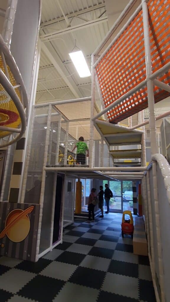 Children and adults explore a multi-level play structure with colorful nets and tunnels, set against a black and white checkered floor, in a well-lit indoor playground.