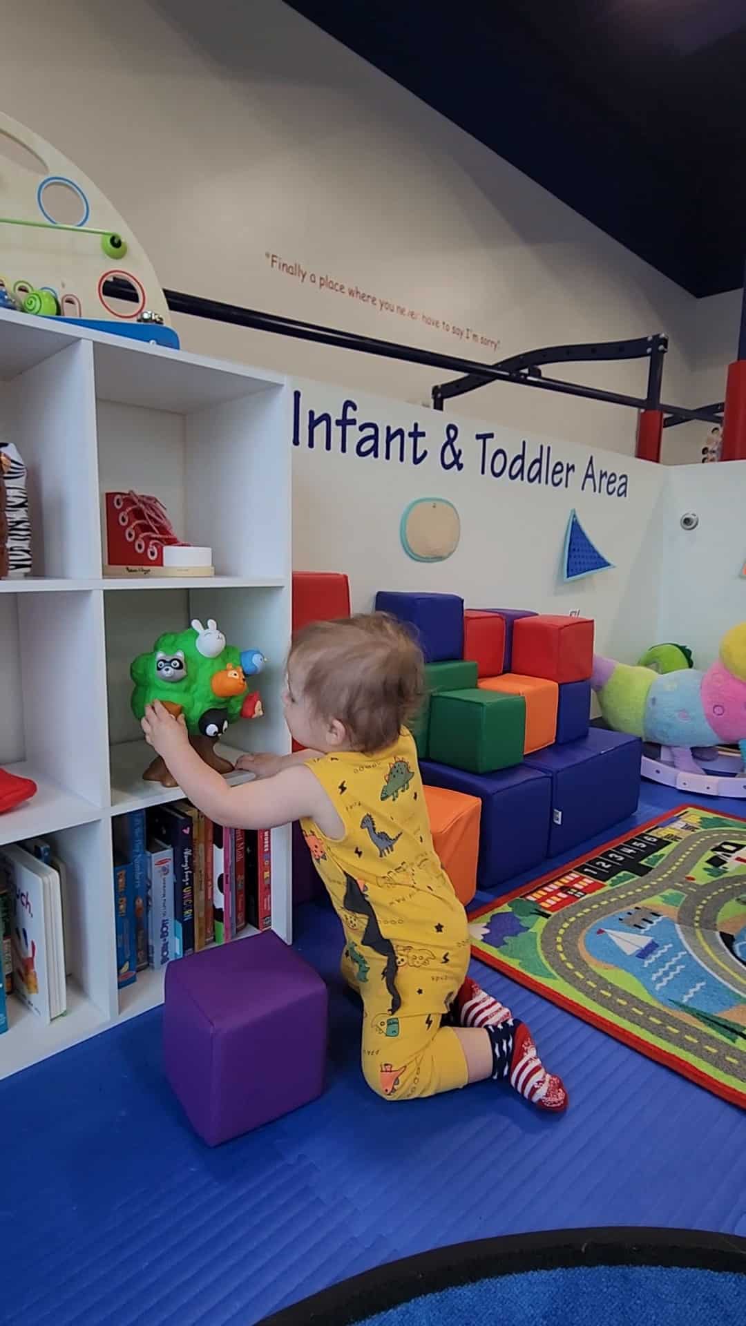 A toddler in a dinosaur-print outfit is selecting toys from a shelf in the Infant & Toddler Area of an indoor play space in Cary, NC, surrounded by colorful soft blocks and a playful rug.