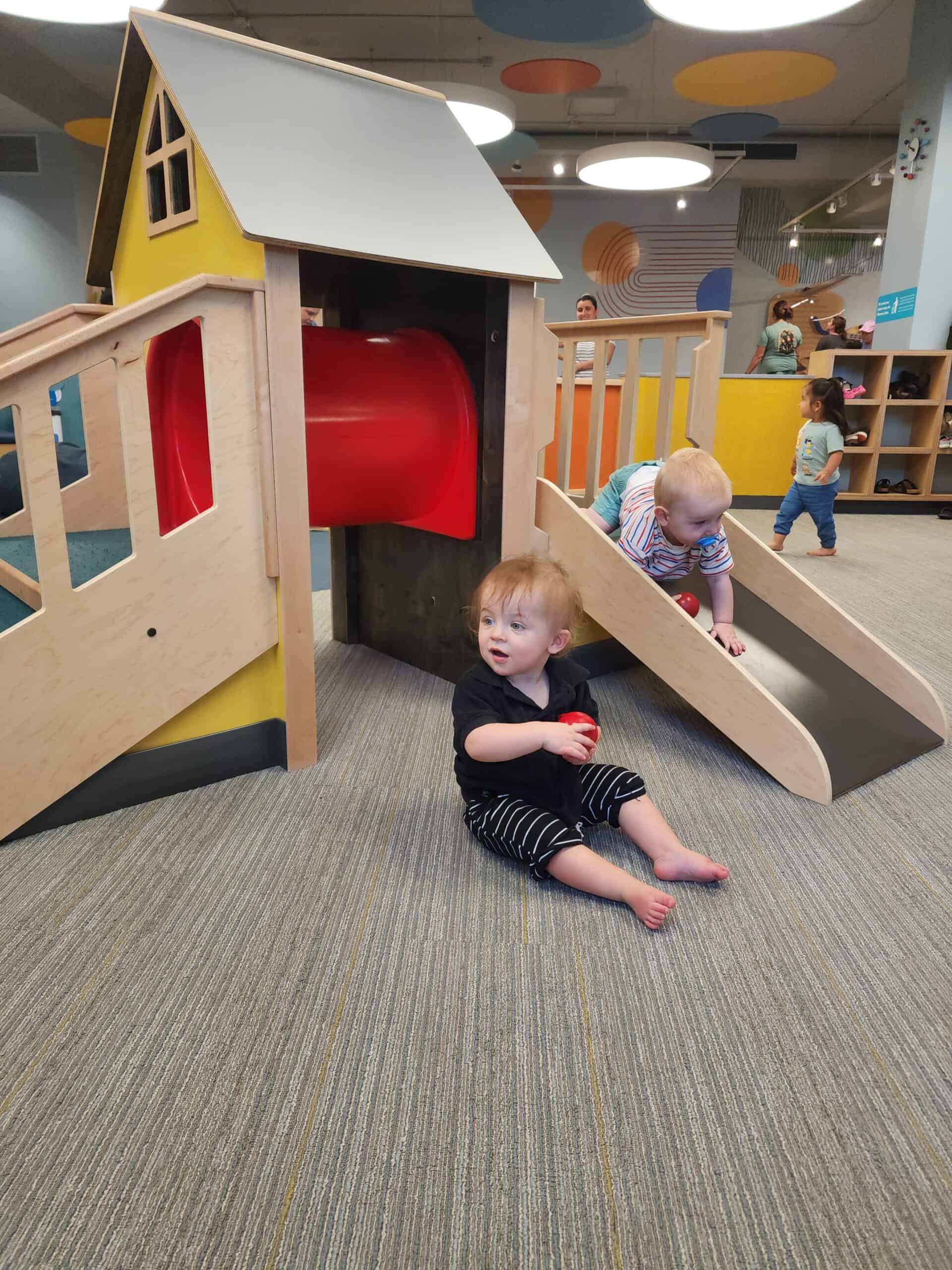 Toddlers enjoy playtime in a cheerful indoor play area, featuring a wooden playhouse with a red slide and a ramp, creating a lively and safe environment for young children to explore and interact