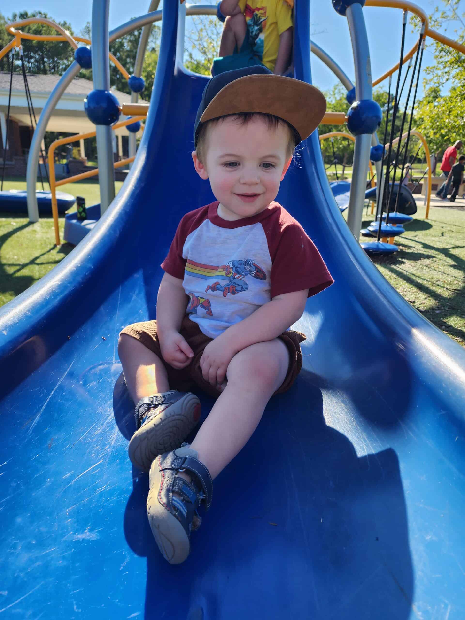 Cheerful toddler with a slight grin, wearing a brown cap, sits on a blue slide at a playground in the Triangle area. His graphic tee and sneakers add a playful touch to this sunny day outdoor activity scene, with other children visible in the background enjoying the equipment.