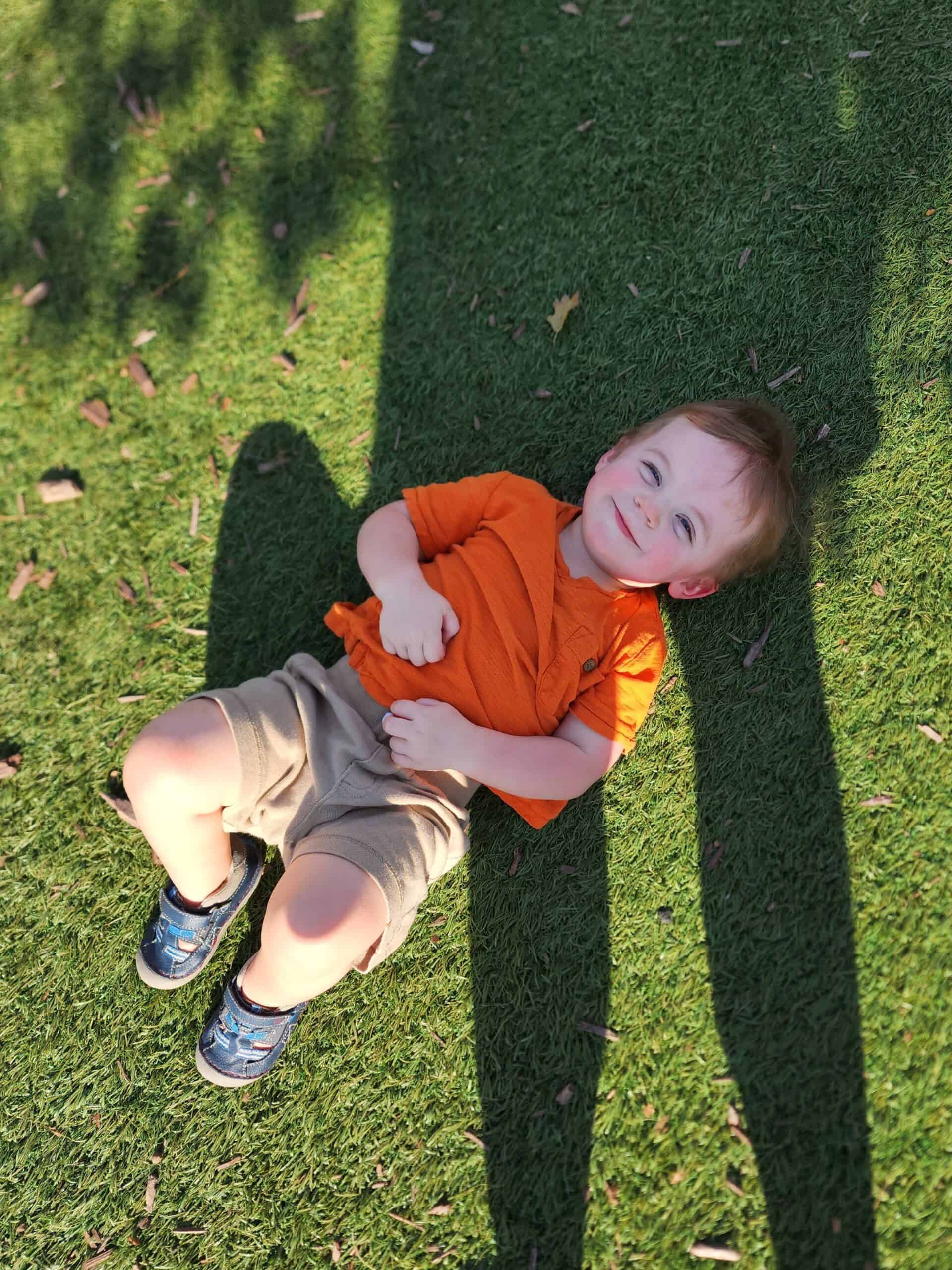 A young child with a beaming smile lies on the grass, wearing an orange shirt and khaki shorts, enjoying the warm sunlight at Northwest Park, Morrisville, NC. The child's shadow stretches out on the grass, capturing a playful and relaxed moment outdoors