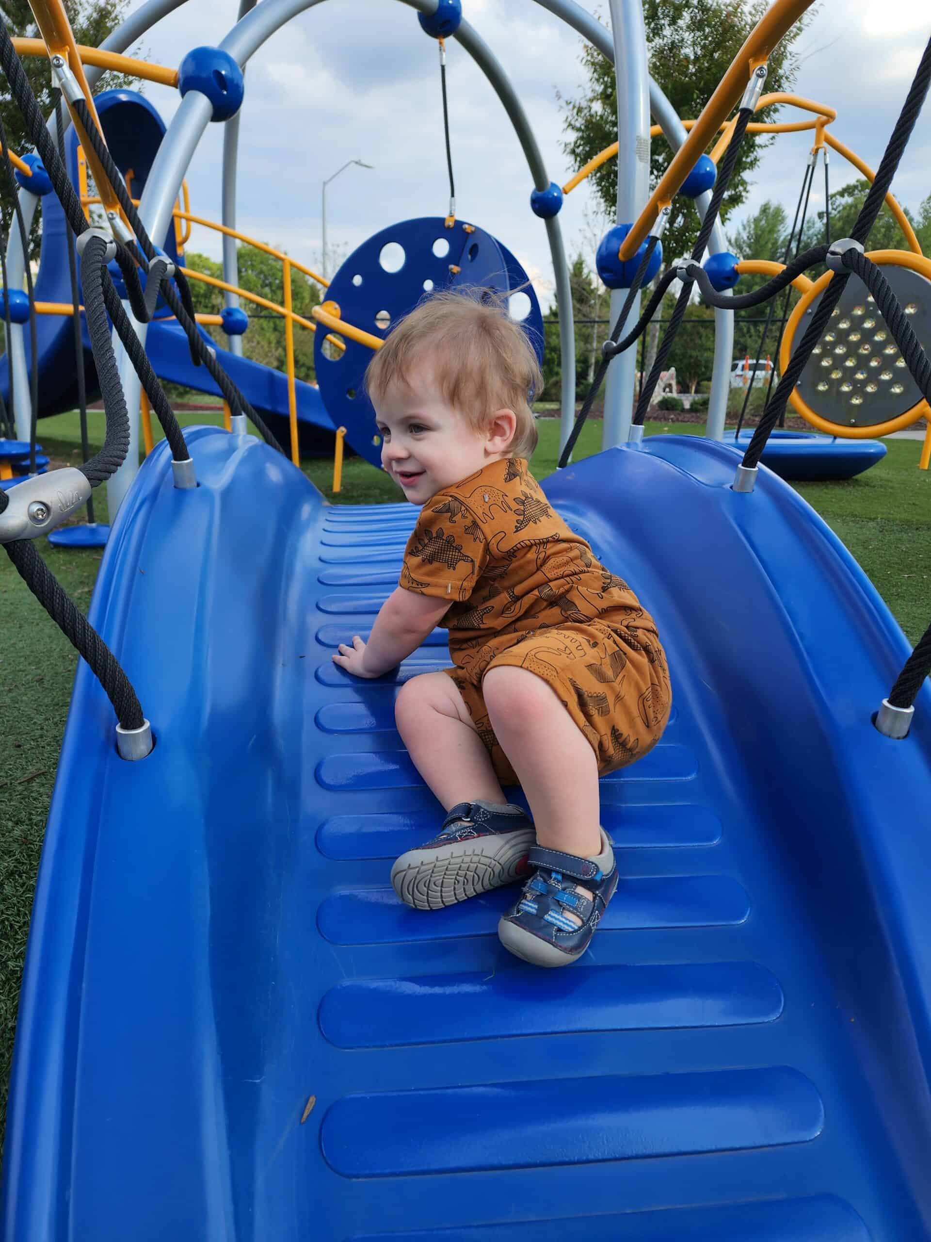 A toddler in a patterned brown outfit sits halfway down a ridged blue slide, looking back with a smile at Northwest Park's playground in Morrisville, North Carolina. The playground's climbing ropes and equipment provide a colorful and engaging background for active play