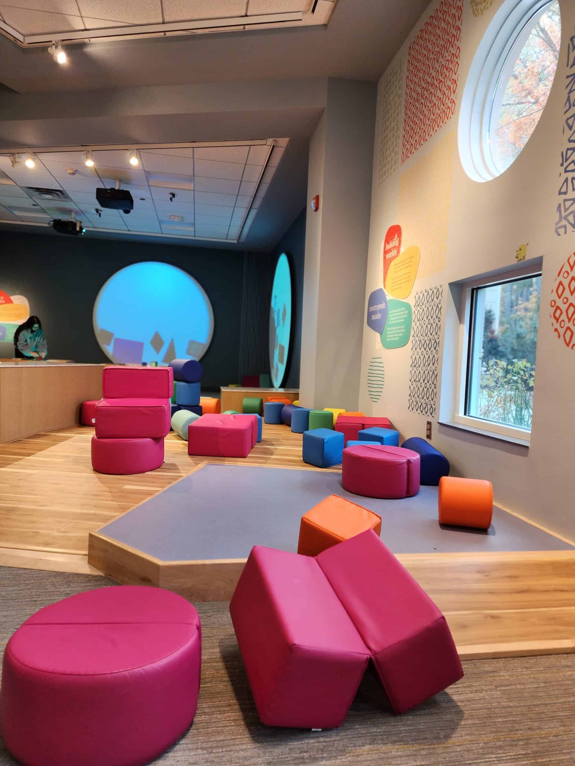 A soft play area for kids with brightly colored foam blocks on a wooden floor, large round windows providing natural light, and educational wall art, creating a safe and inviting space for imaginative play and motor skill development.