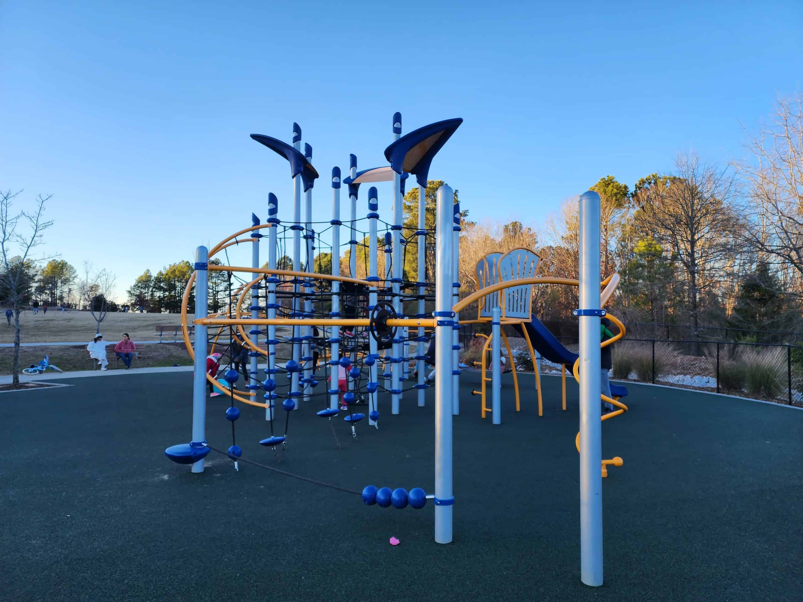 A vibrant playground at Northwest Park, Morrisville, NC, with children playing in the background. The main structure features blue and yellow climbing equipment, including slides and interactive play panels. The playground's rubber surface ensures safety, and the surrounding open grassy areas invite active play in a community-friendly environment.