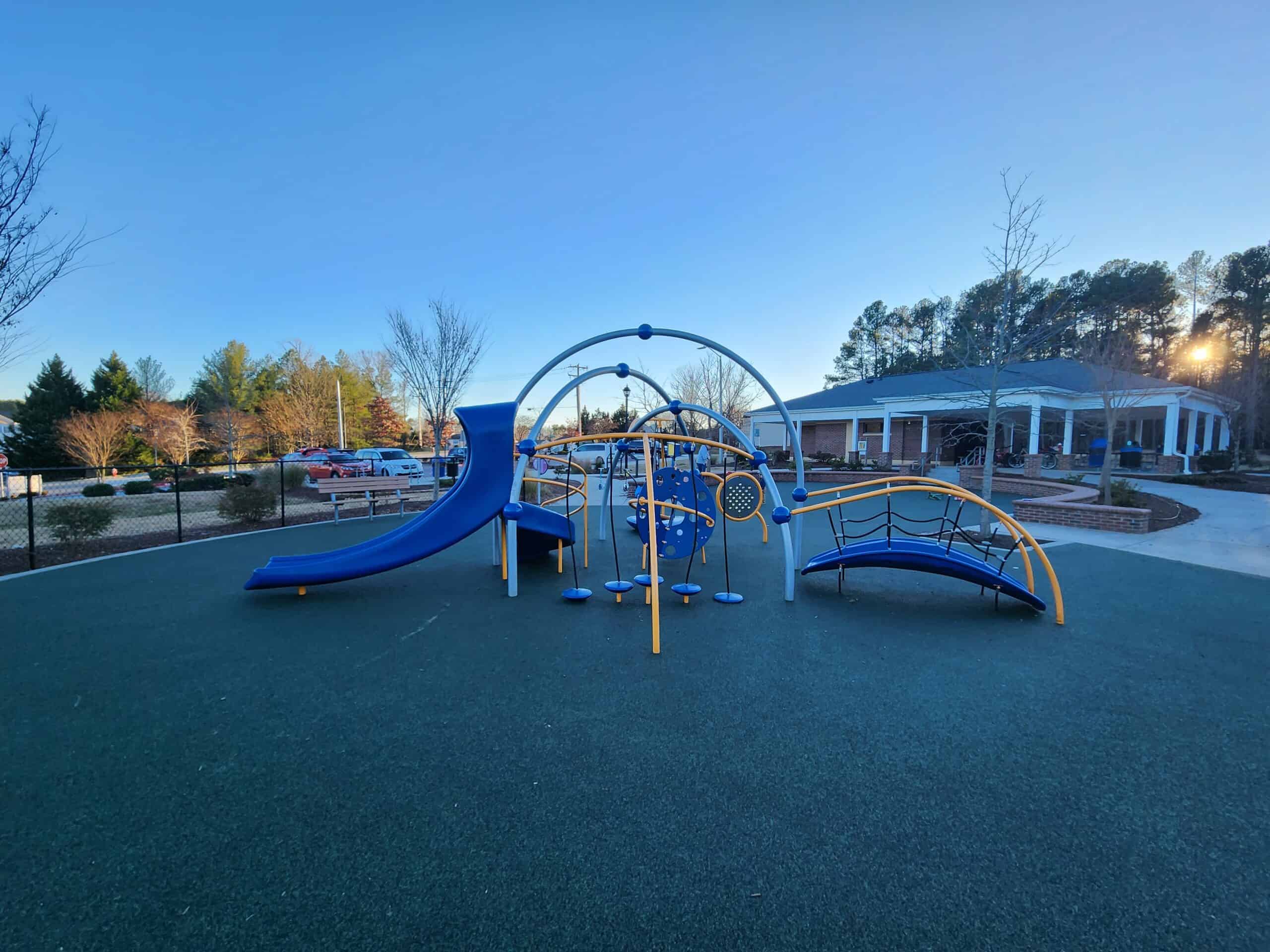 Northwest Park in Morrisville, NC, featuring a playground with blue and yellow equipment. The image shows a curved blue slide, overhead climbing apparatus, and a low-to-ground balance structure on a green rubber surface. The sun is setting behind a building with white columns, giving a serene atmosphere to the park