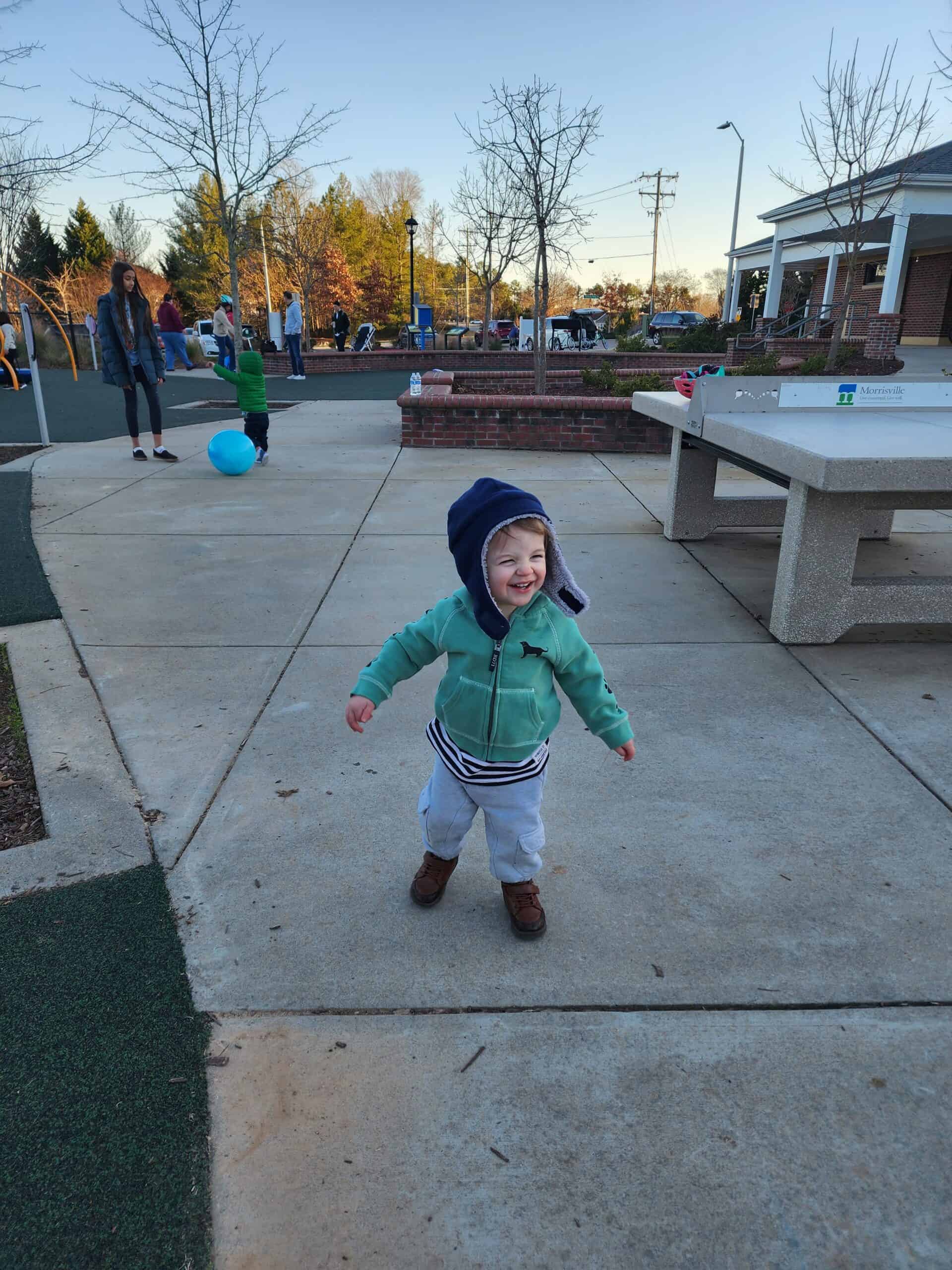 Excited toddler in a green hoodie and blue beanie walking towards the camera on a concrete path at a playground in Morrisville, North Carolina, with joyful expression. The background includes park benches and visitors enjoying the outdoor setting.