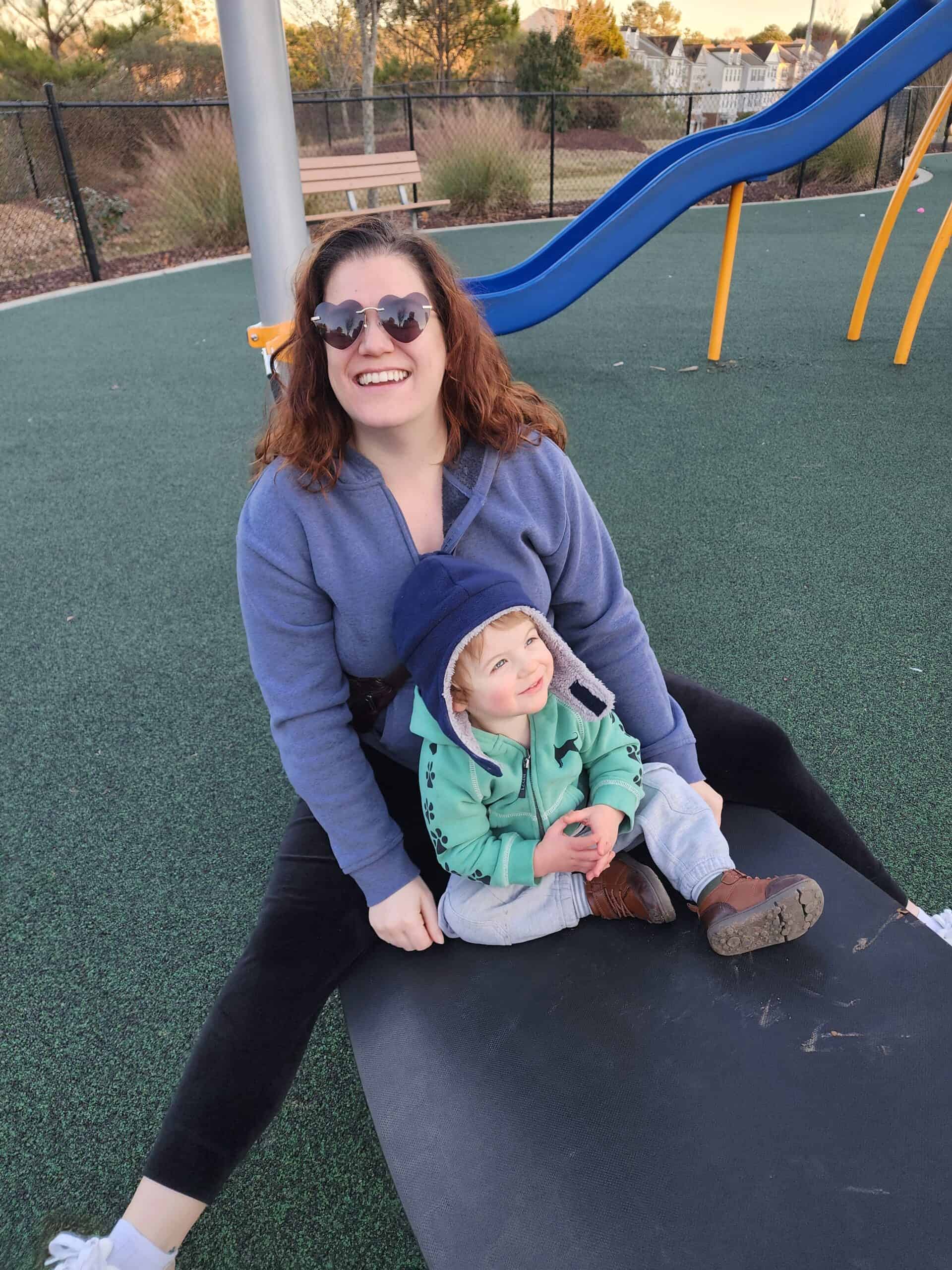 A joyful moment as a woman with heart-shaped sunglasses smiles broadly, seated on a playground mat with a young child on her lap. The child, wearing a green jacket and a beanie, looks up with a delighted expression. In the background, there's a blue slide and a bench, indicative of a well-maintained park setting during what appears to be dusk.