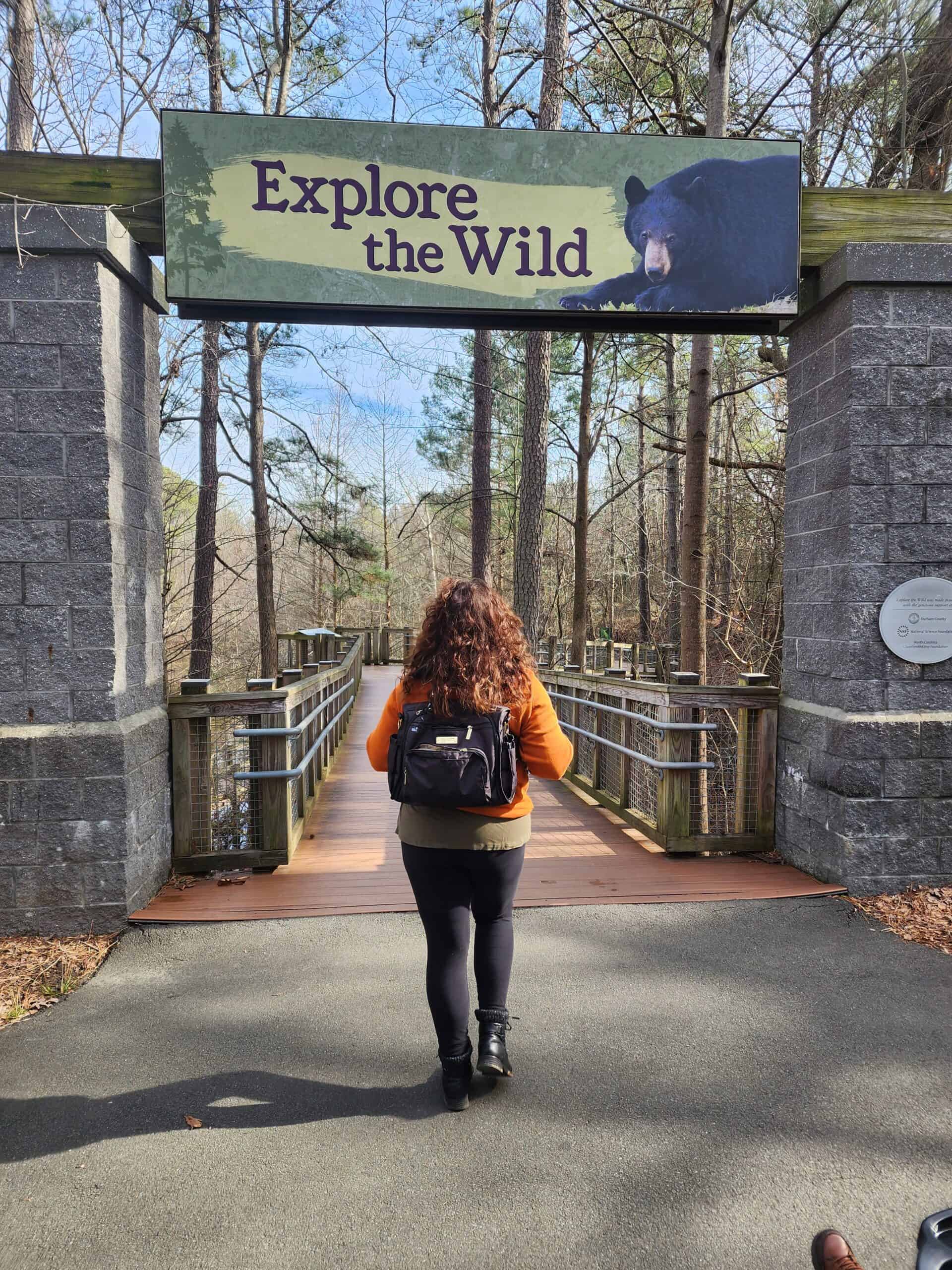 A visitor with a backpack approaches the 'Explore the Wild' entrance at the Museum of Life and Science in Durham, framed by a serene woodland setting and a welcoming sign featuring a black bear image.