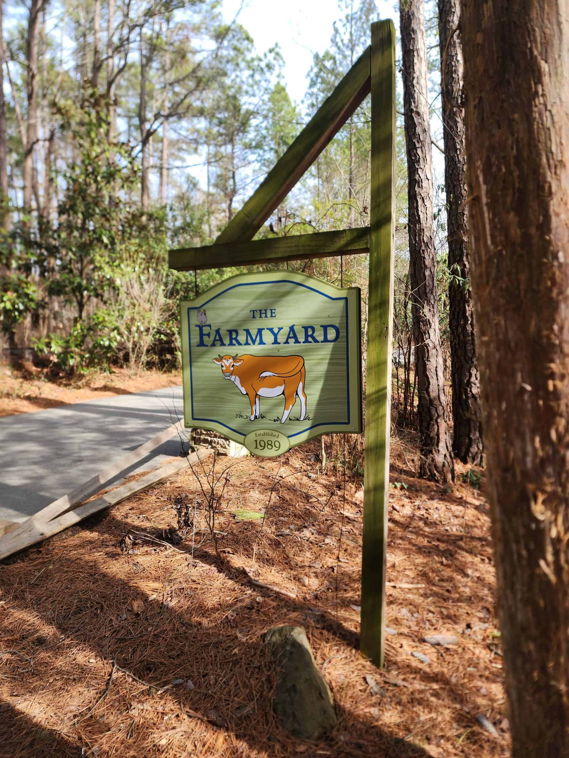 Signpost for 'The Farmyard' set amidst a backdrop of tall pines and a carpet of pine needles, marking a pastoral exhibit established in 1989, suggesting an educational farm experience within a natural forest setting