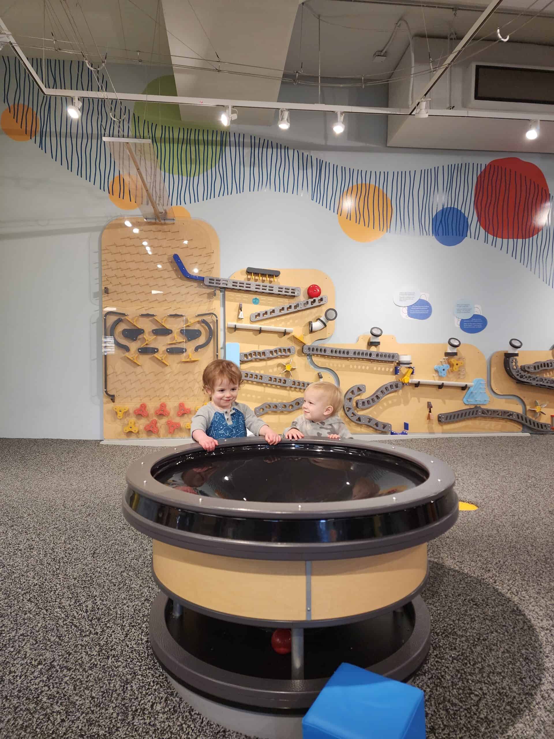 Two young children play together at a circular activity table in a Durham, NC, venue, which is one of the fun and educational things to do with kids in the area. The background is adorned with an interactive wall display featuring ball tracks and colorful patterns