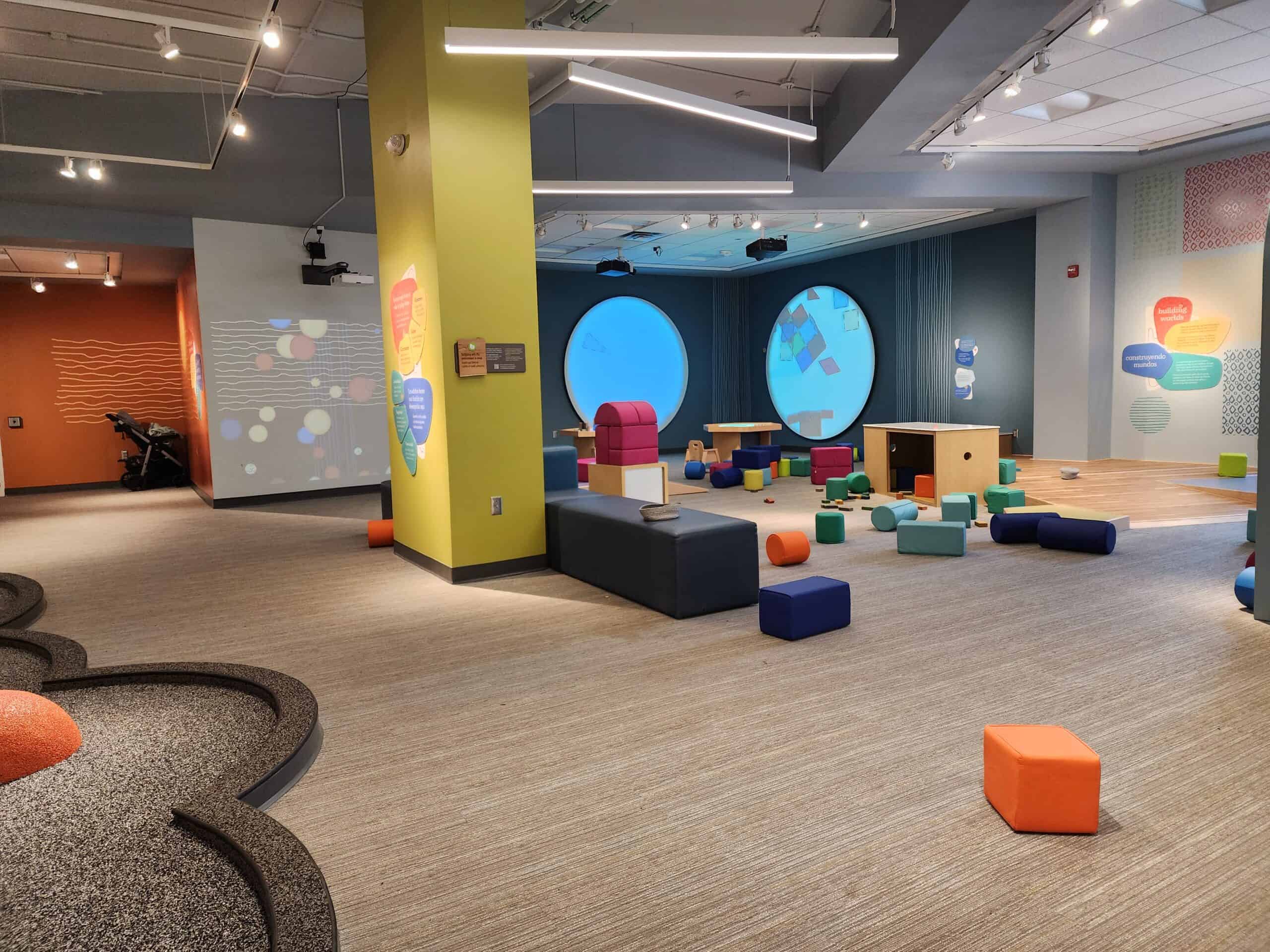 Spacious and modern children's playroom with colorful foam blocks scattered across the floor, large circular windows, and educational wall graphics, providing a bright and inviting space for interactive learning and play.