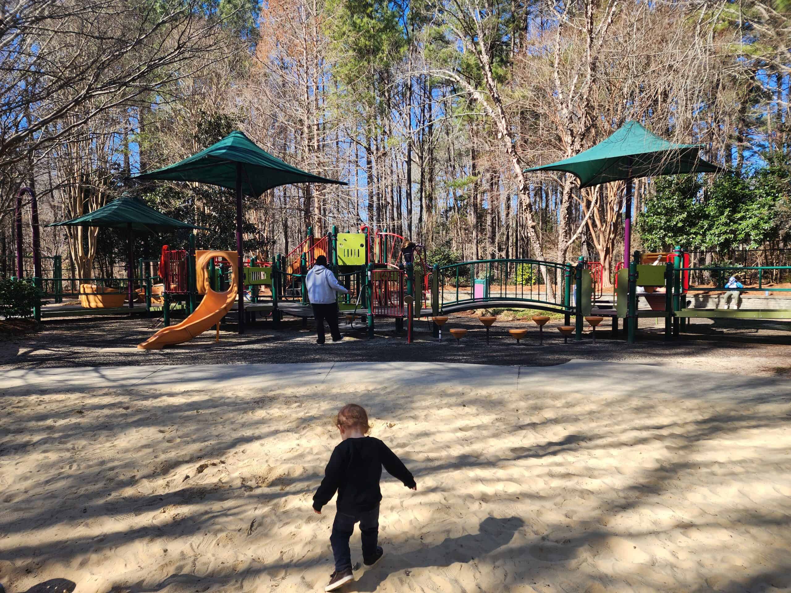A child runs towards the inviting play area at a sunny playground in Cary, North Carolina, surrounded by tall pine trees. The playground features various equipment with shades of green canopies, sand-covered ground, and is filled with the energy of children's playtime activities.