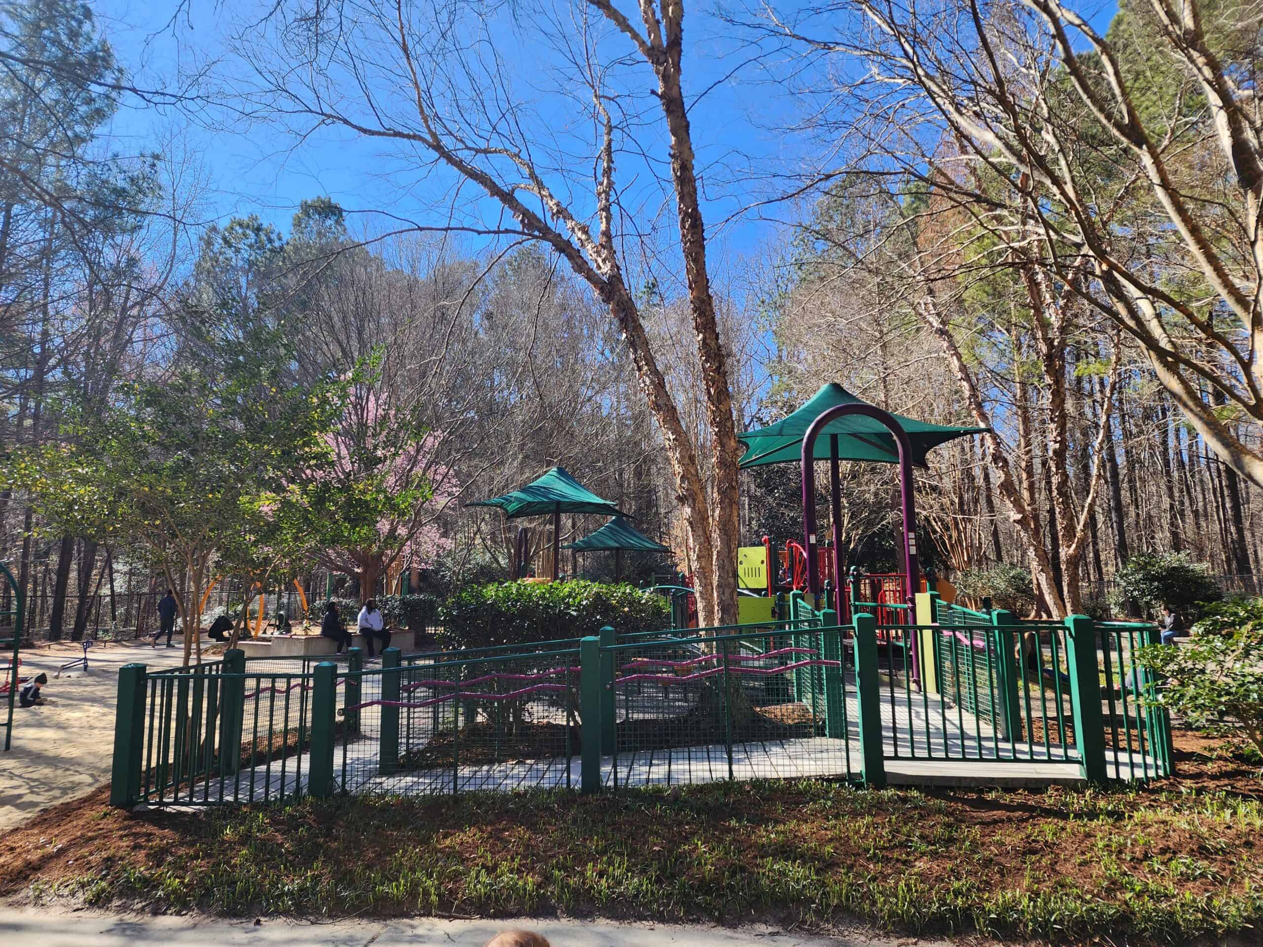 A scenic view of a playground in Cary, NC, under a clear blue sky. The park is framed by lush trees and blooming pink flowers, with children enjoying the green and red play structures. The fenced perimeter ensures a safe play environment, highlighting the natural and well-maintained setting.