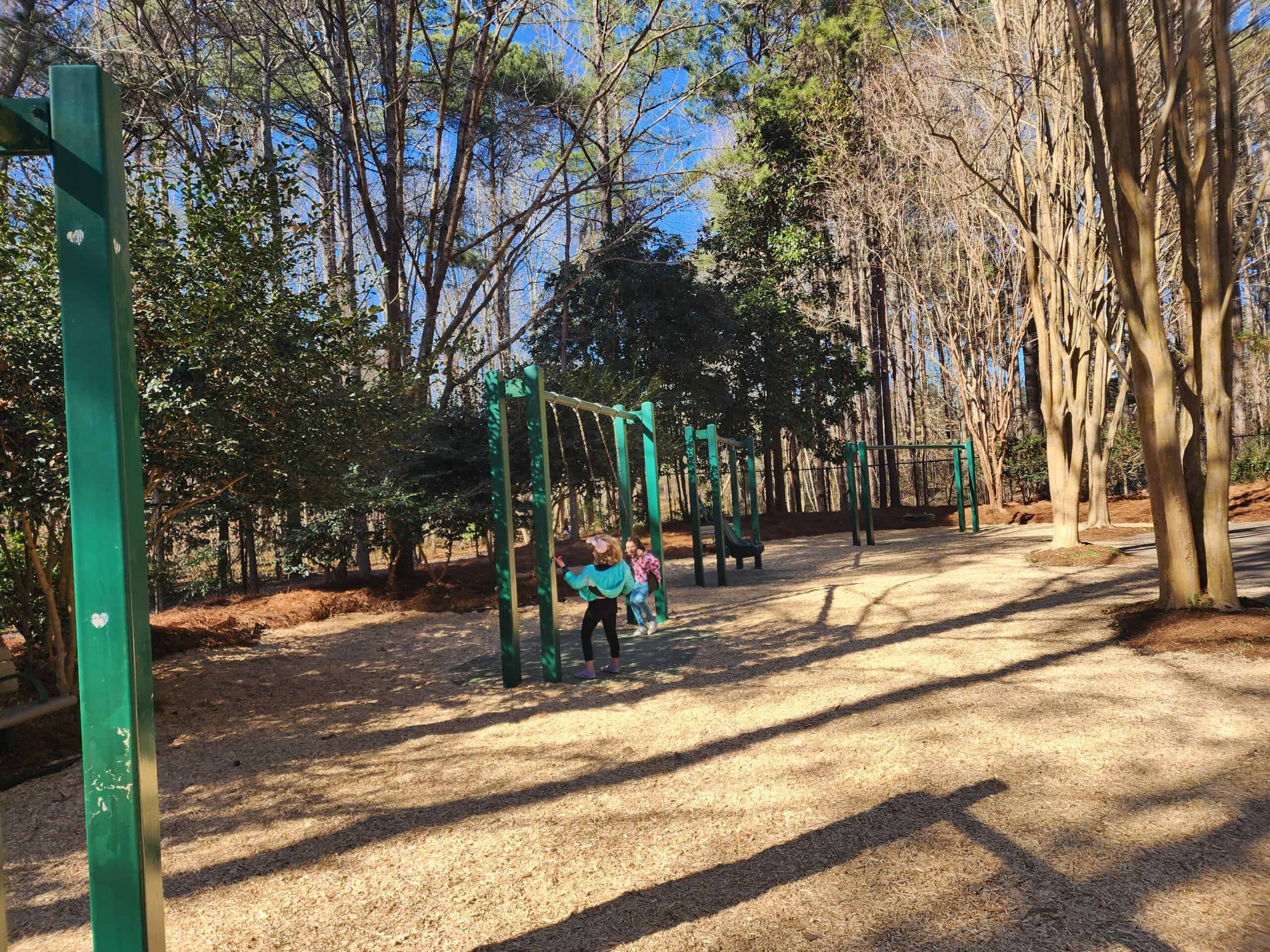 Children play on green swing sets at a Cary, NC, playground nestled among tall, slender trees casting long shadows on the mulch-covered ground. The serene park atmosphere is infused with the energy of childhood joy and the tranquility of a forest setting