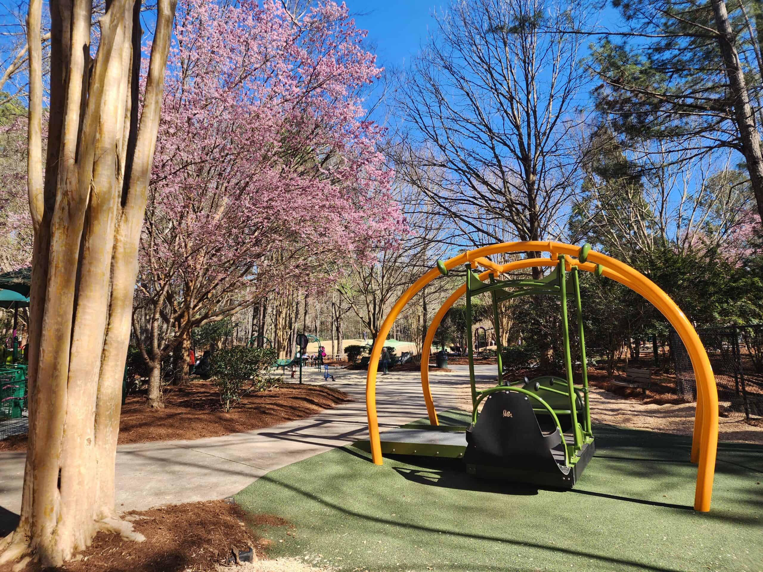 An accessible, inclusive swing set awaits its next visitor at a Cary, NC playground. Surrounded by a vibrant landscape with blooming pink cherry blossoms and the contrasting bare branches of winter trees, this park provides a welcoming environment for children of all abilities against a bright blue sky.