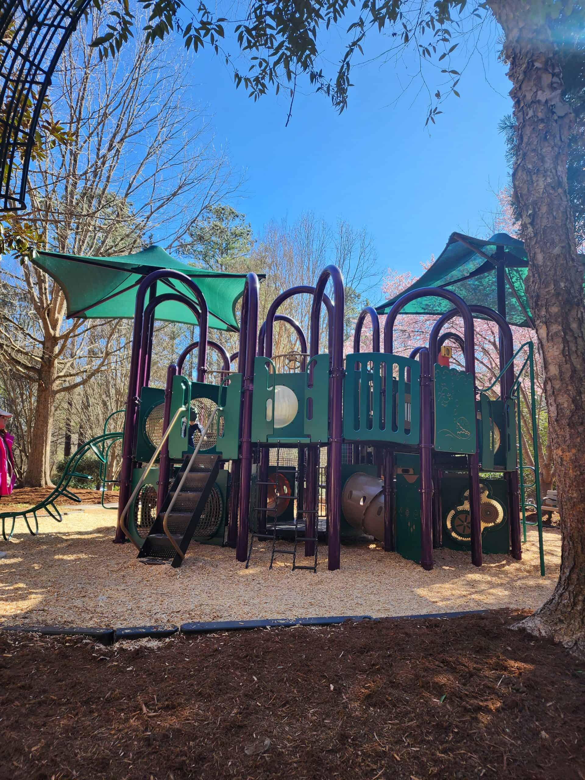a multi-level playground structure with green shades and purple railings stands amidst a natural setting with mature trees and a mulch-covered ground. The playground invites children to climb, slide, and explore in the heart of Cary, North Carolina