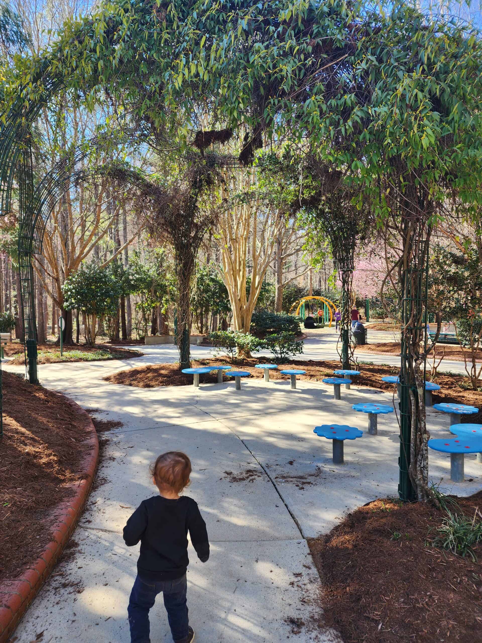 A young child approaches a whimsical outdoor area with blue mushroom-shaped stools under the shade of an arching trellis covered in greenery, in a peaceful park setting in Cary, North Carolina. The path, bordered with red edging, leads to an inviting playground in the distance, nestled among tall trees and a clear blue sky