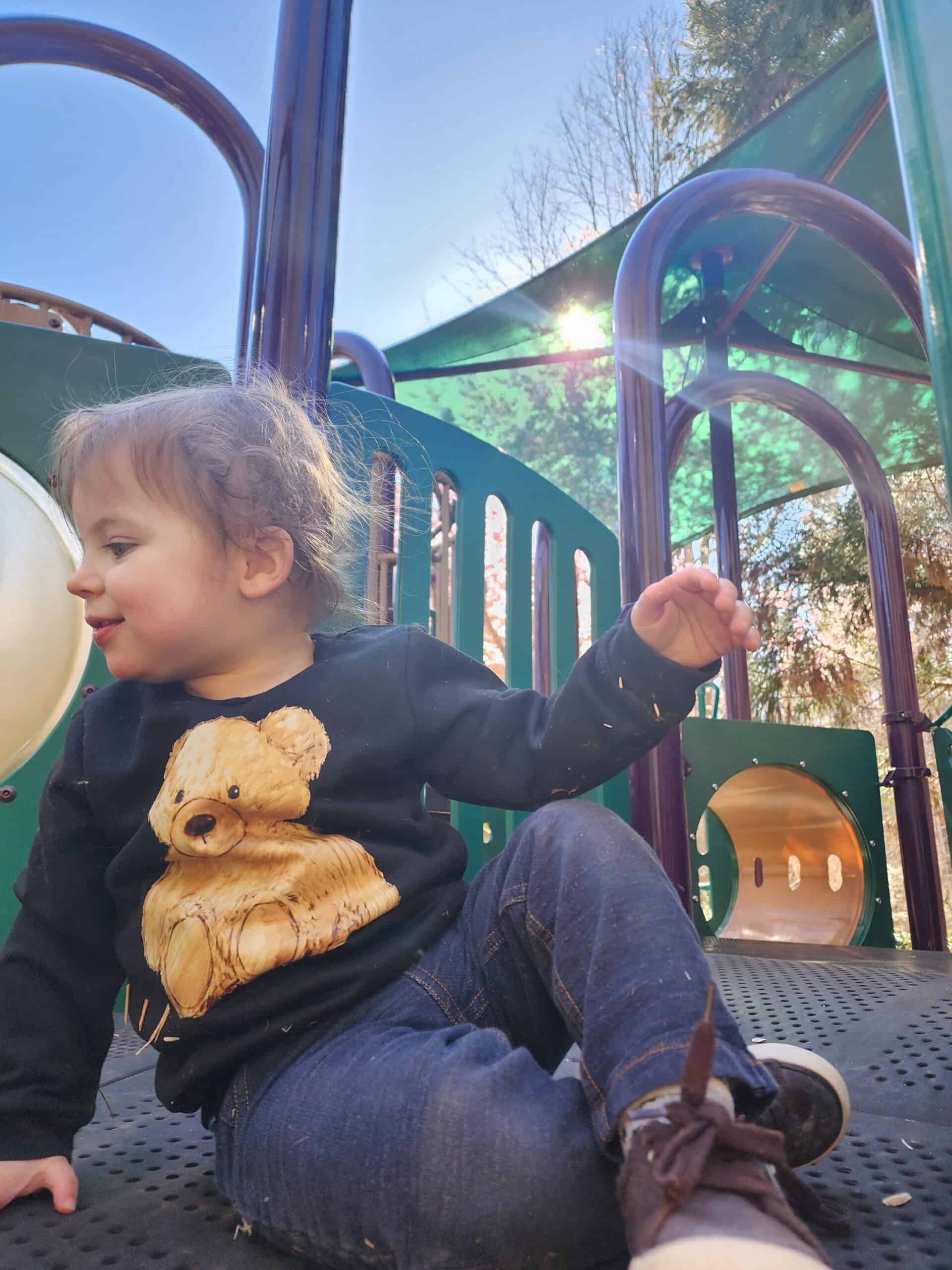 A young child with a joyful expression sits on a playground in Cary, North Carolina, with rays of sunlight filtering through the equipment. The child wears a black sweater adorned with a teddy bear graphic and blue jeans, capturing a moment of innocent playtime.