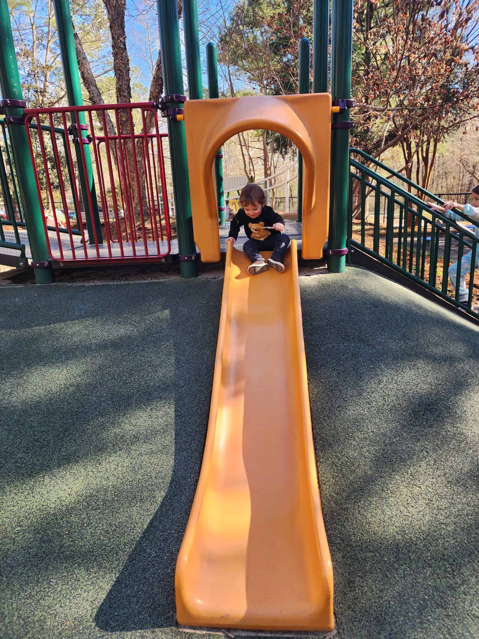 A delighted child with a teddy bear on their sweater pauses at the top of a bright orange slide, ready for fun at a Cary, North Carolina playground. The sturdy green railings and surrounding trees create a safe and natural play environment on a sunny day