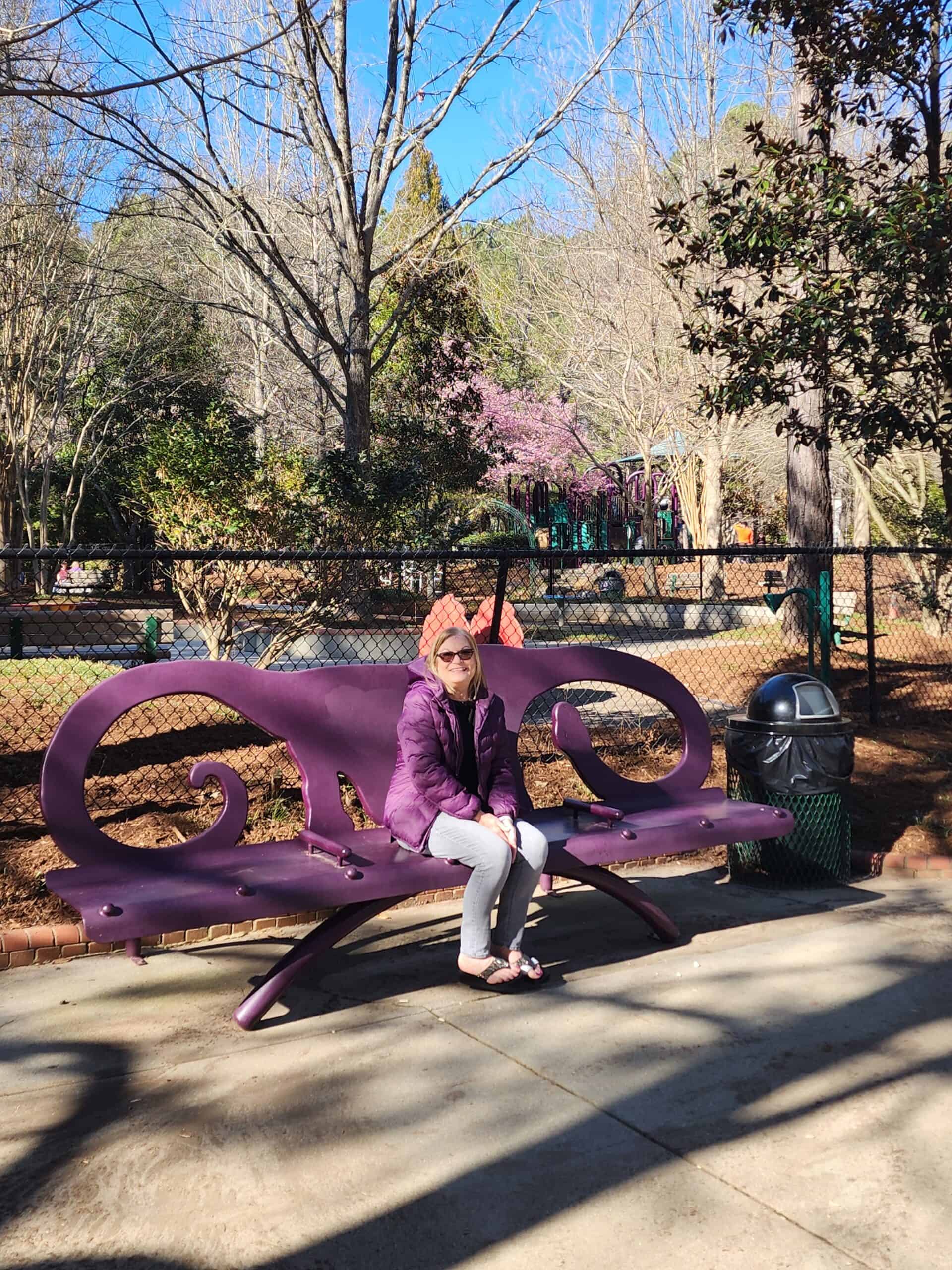 A person sits on a unique, ornate purple bench at a playground in Cary, North Carolina, with a backdrop of a fenced playground area, blooming pink cherry blossoms, and leafless trees. The scene evokes a leisurely and peaceful day at the park, with the bench's design adding a touch of whimsy to the setting