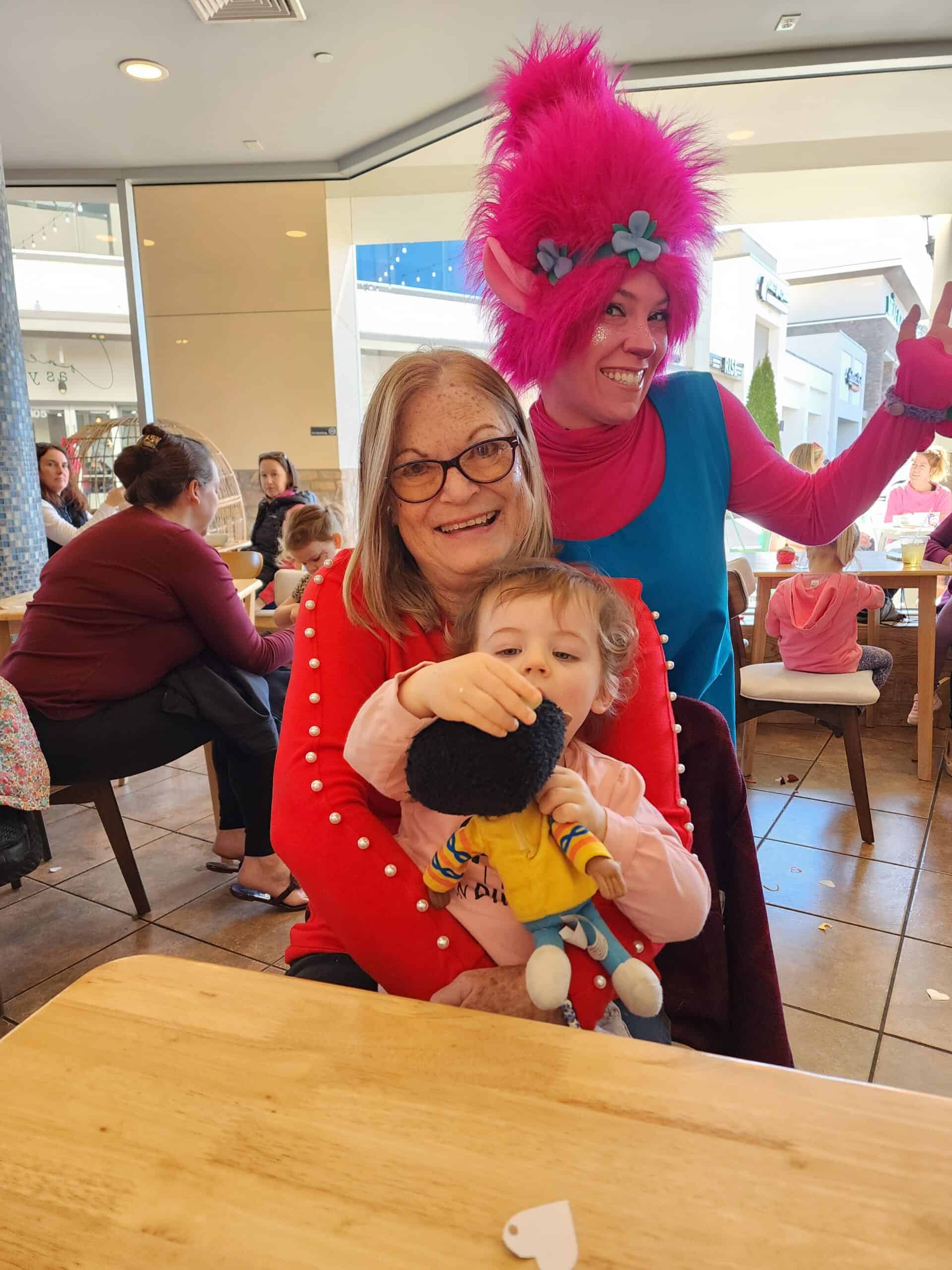A joyful multi-generational family moment at a toddler activity in Cary, NC, with a senior woman smiling at the camera, a child engaging with a toy, and a vibrant character with pink hair and costume spreading cheer.