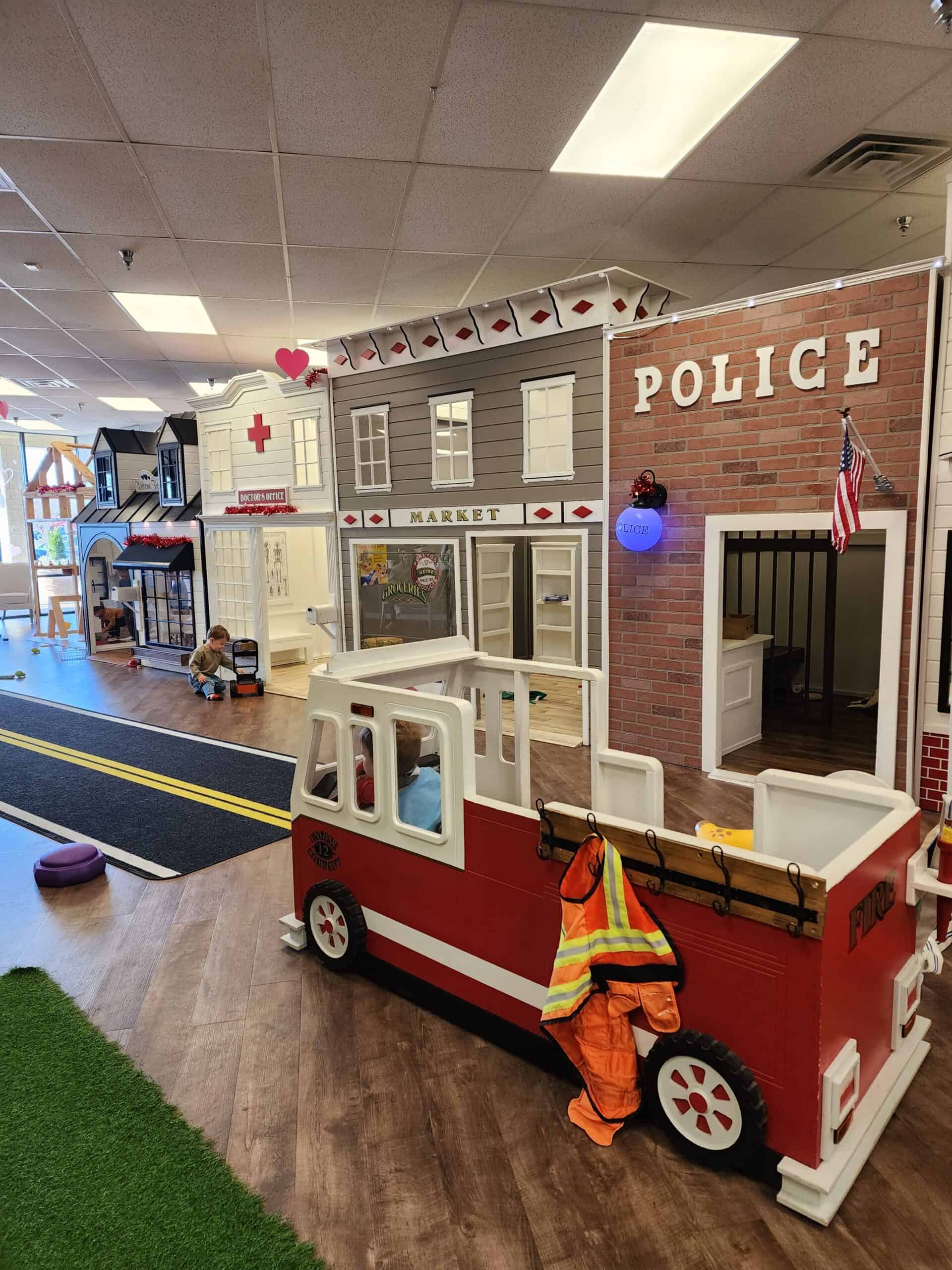 A playful and educational setup for children in an indoor play area, featuring a mini-town with a fire station, police station, and market, complete with a toy fire truck and realistic costumes for imaginative toddler activities.