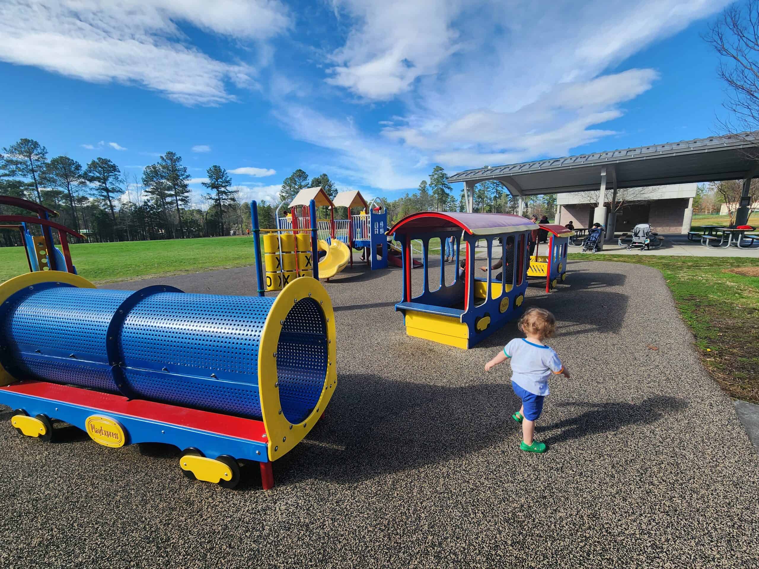 A young child toddles towards colorful playground equipment on a sunny day, with a vibrant blue tunnel and a yellow and red play train set against a background of green grass and a clear sky.