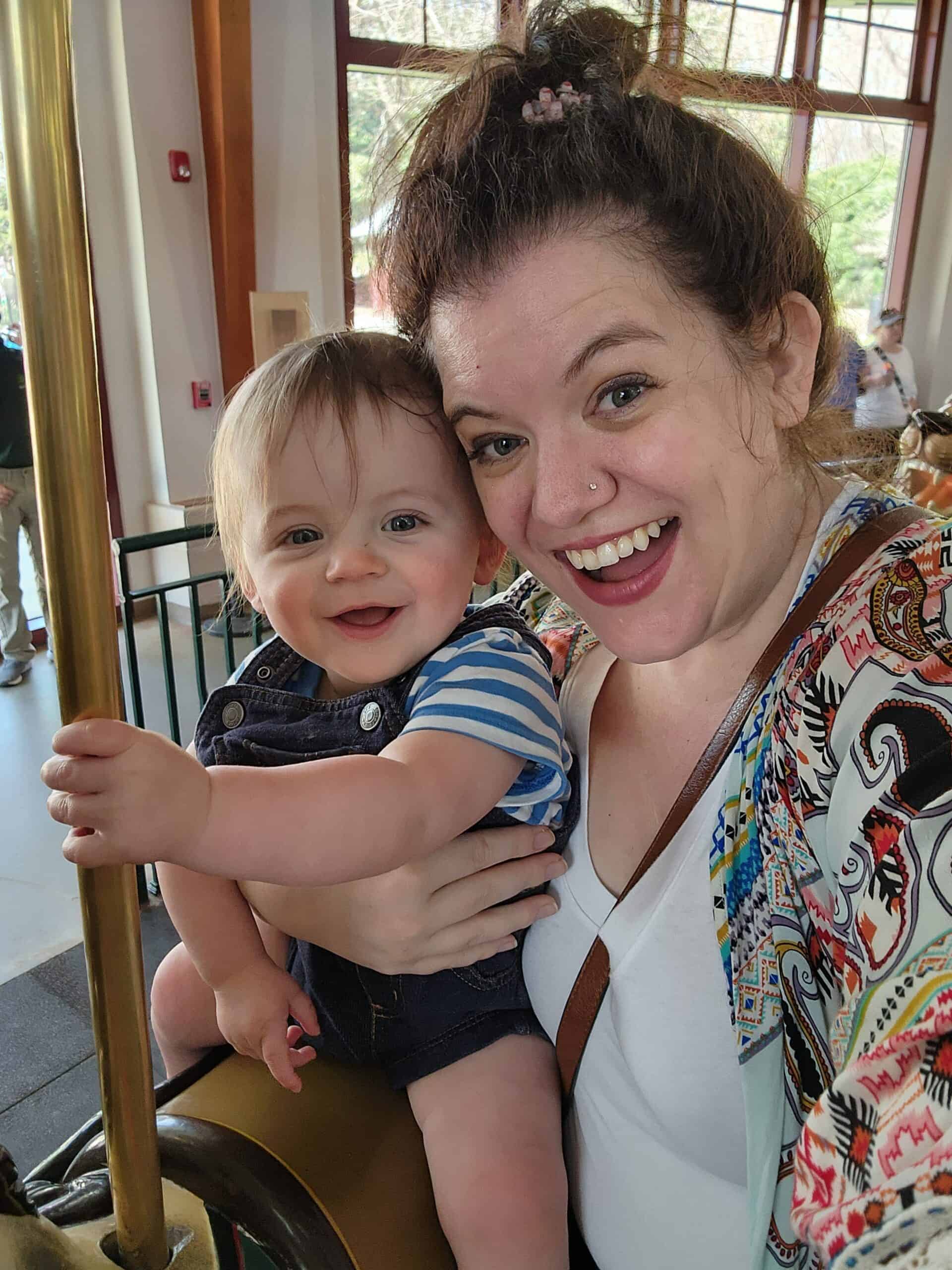 A heartwarming selfie of a woman and a baby sharing a joyful moment on a carousel ride, with the baby gripping the golden pole and both wearing bright, happy smiles