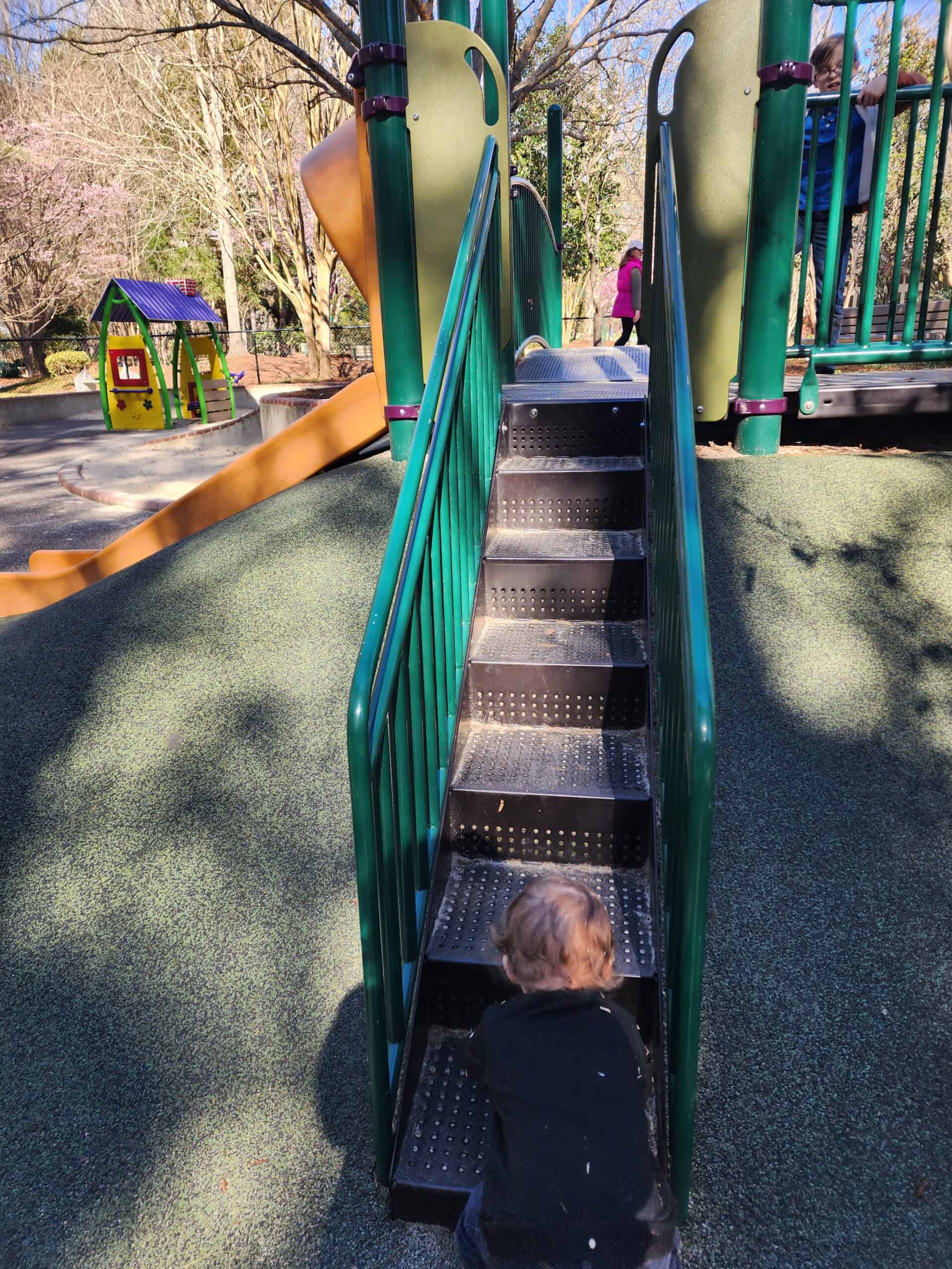 A young child in a black sweater climbs the stairs of a playground slide, with the early springtime sun casting long shadows on the rubber floor, and a glimpse of pink blossoms in the distance.