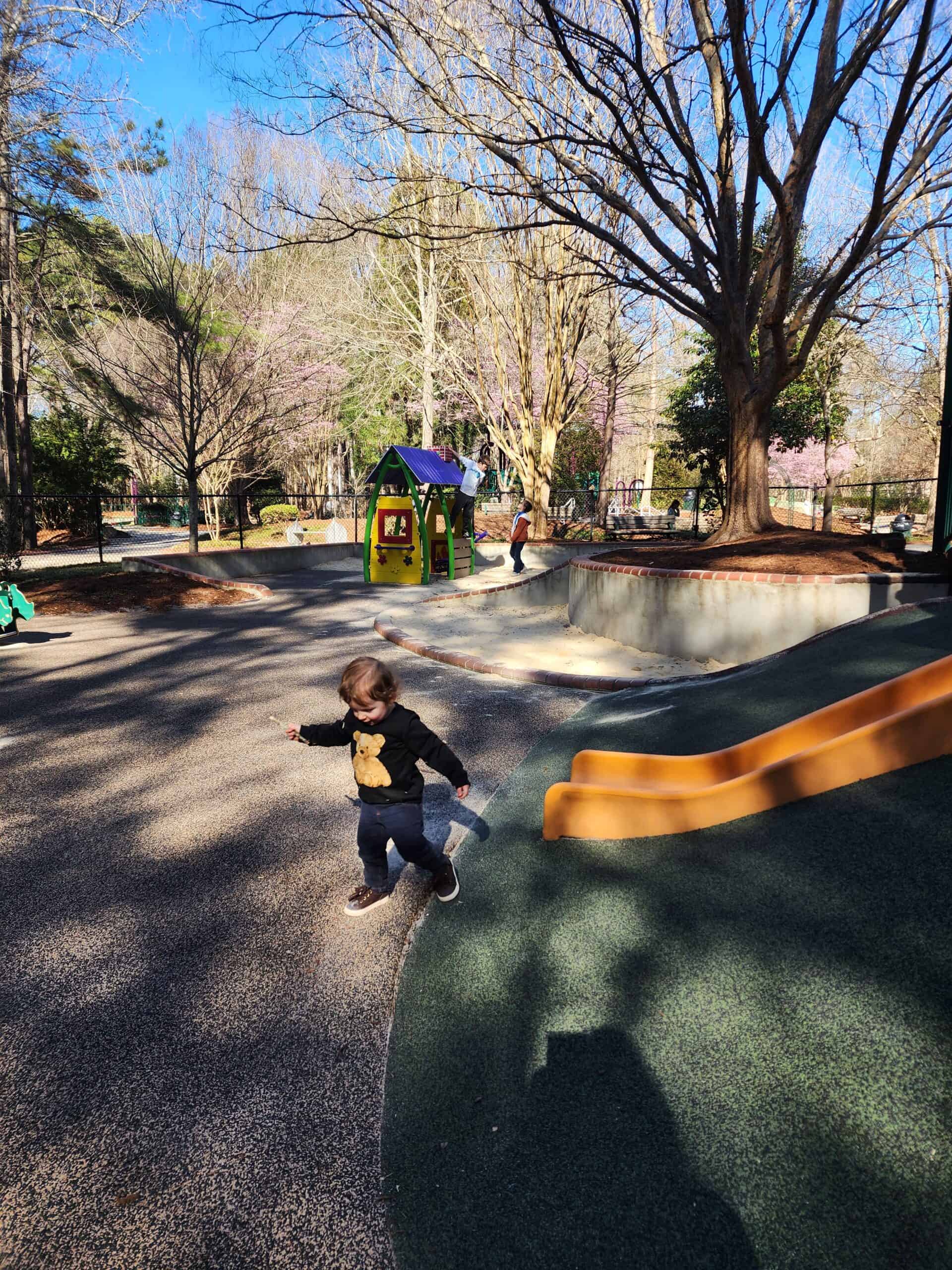 A young child runs joyfully on a shadow-dappled playground in Cary, NC, with spring blossoms visible on trees in the background, capturing a moment of carefree play