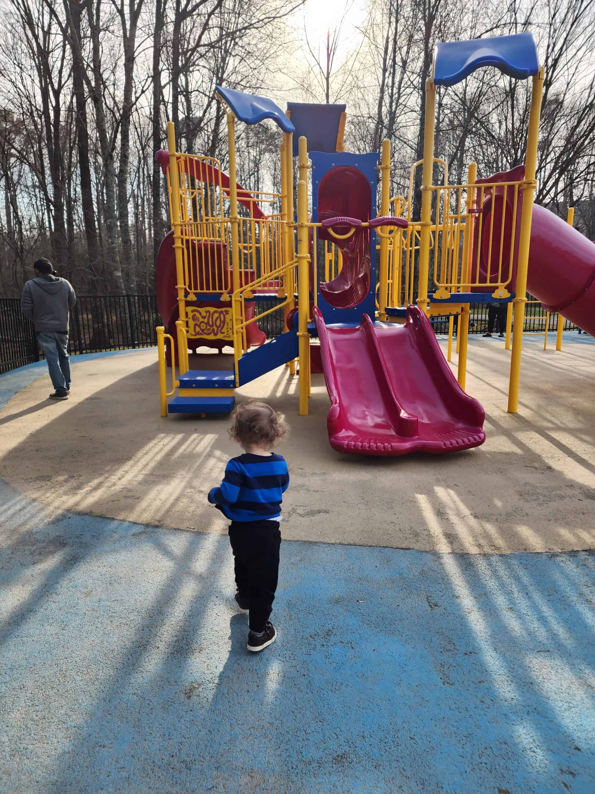 A curious toddler in a blue and black striped shirt walks towards a colorful playground with yellow railings and a vibrant red slide, with bare trees in the background and a blue safety surface underfoot.