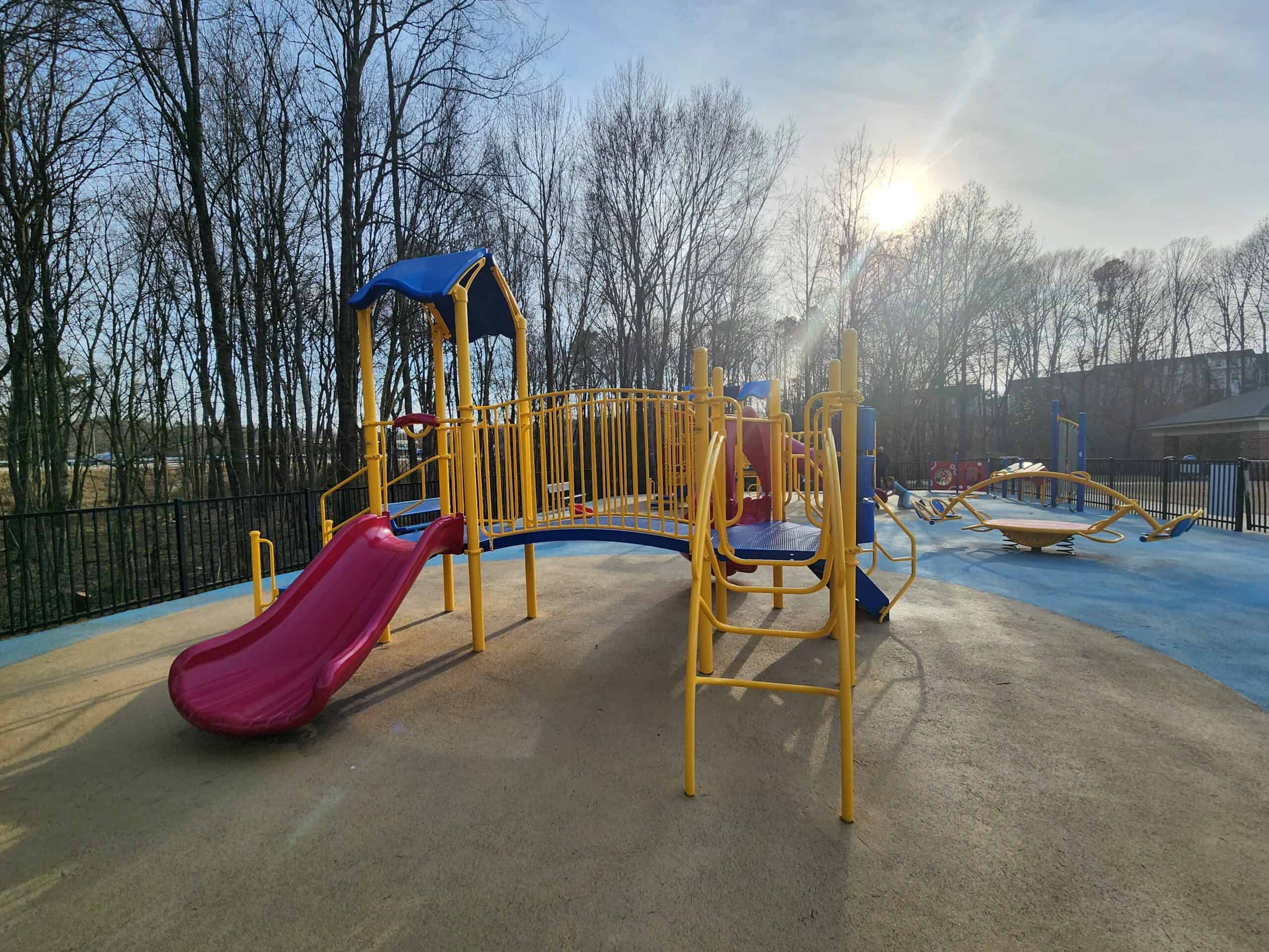 The warm glow of the setting sun illuminates a vibrant playground featuring yellow and blue equipment, including slides and climbing structures, set against a backdrop of leafless trees and a clear sky.