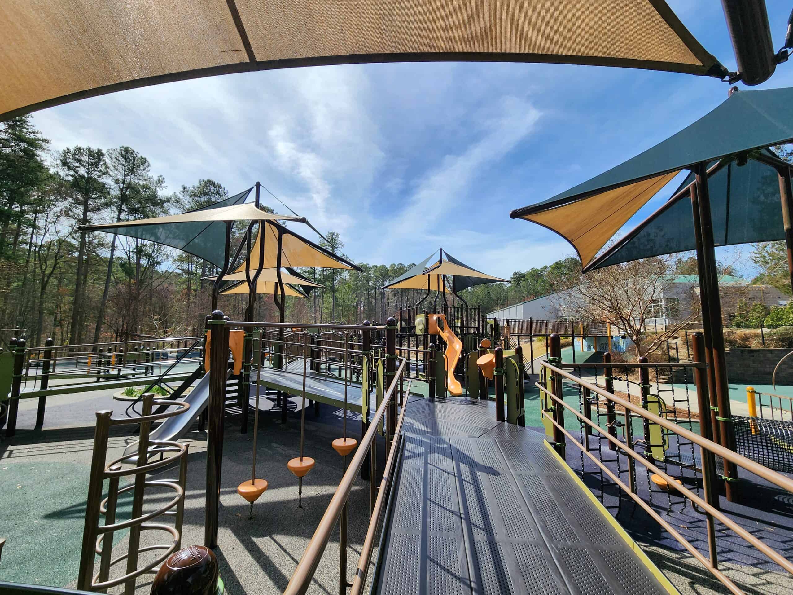 A modern playground bathed in sunlight with protective canopies casting cool shade over various climbing and play structures, set amidst a tranquil forest setting.