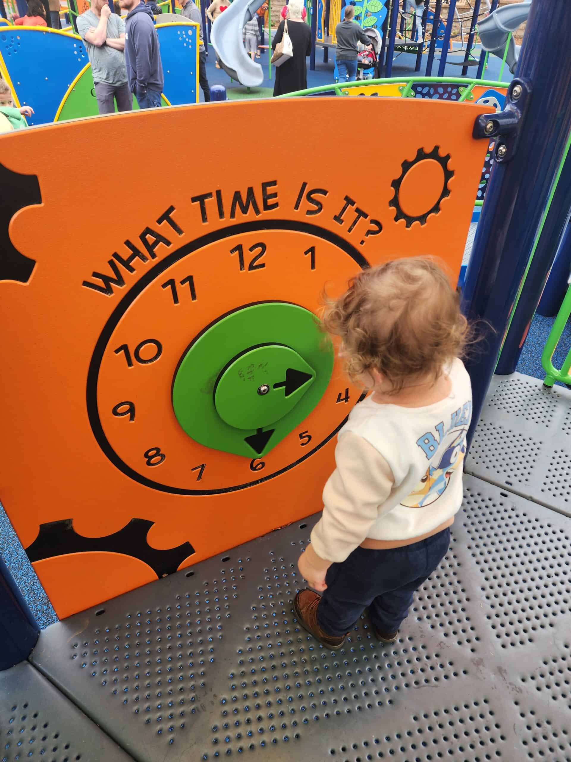 A curious toddler at Holding Park playground in Wake Forest examines a colorful learning panel with a large 'What Time Is It?' clock in shades of orange and green - an example of what makes this a toddler-friendly playground in the Triangle area 