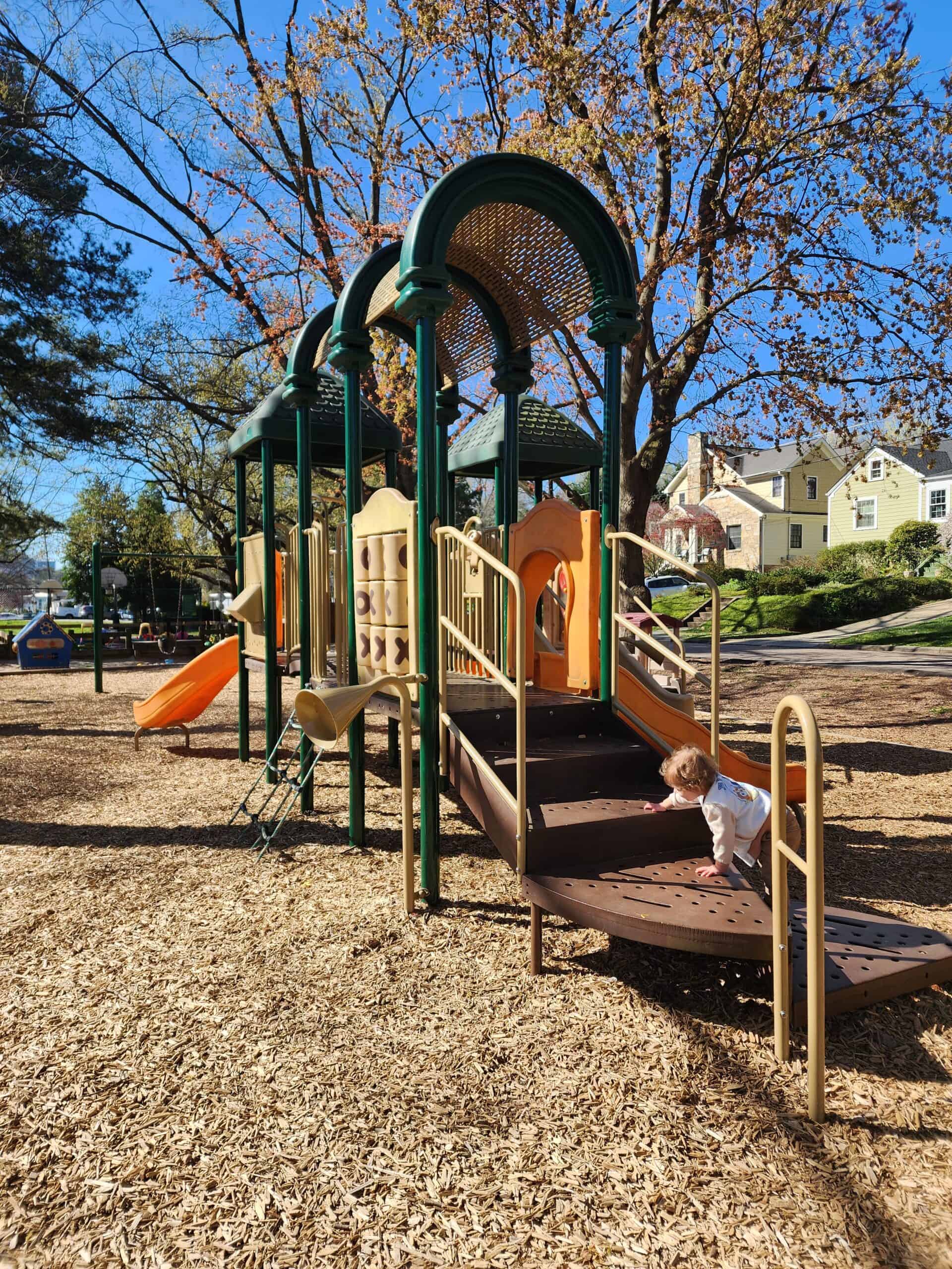 A child climbs the stairs of a colorful playground set with slides and climbing structures on a sunny day, surrounded by wood chips and with residential houses and autumn trees in the background