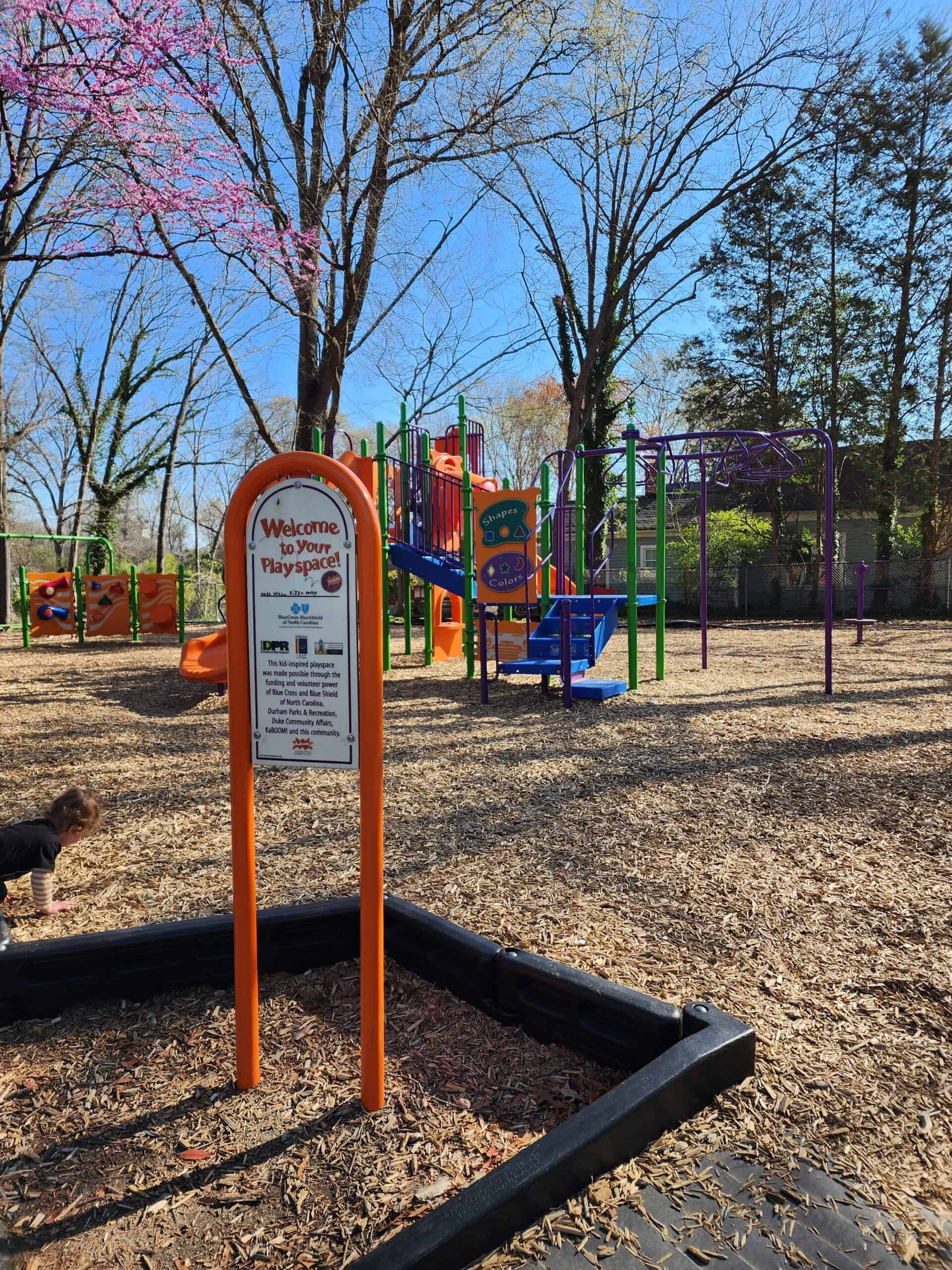 The entrance to Burch Avenue Playground is marked by a vibrant orange sign that reads 'Welcome to Your Playspace!' with a colorful, multi-structure playground in the background. The scene is lively with children playing, a blossoming pink tree, and a clear blue sky.