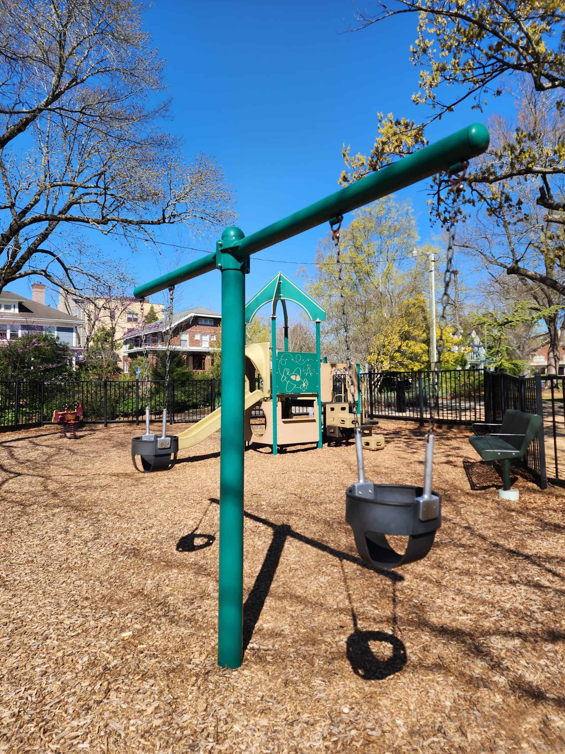 Foreground shows a close-up of a swing set with black rubber swings hanging from a green metal frame, with a playground structure in the background featuring slides and interactive elements, all against the backdrop of a clear blue sky and leafless trees