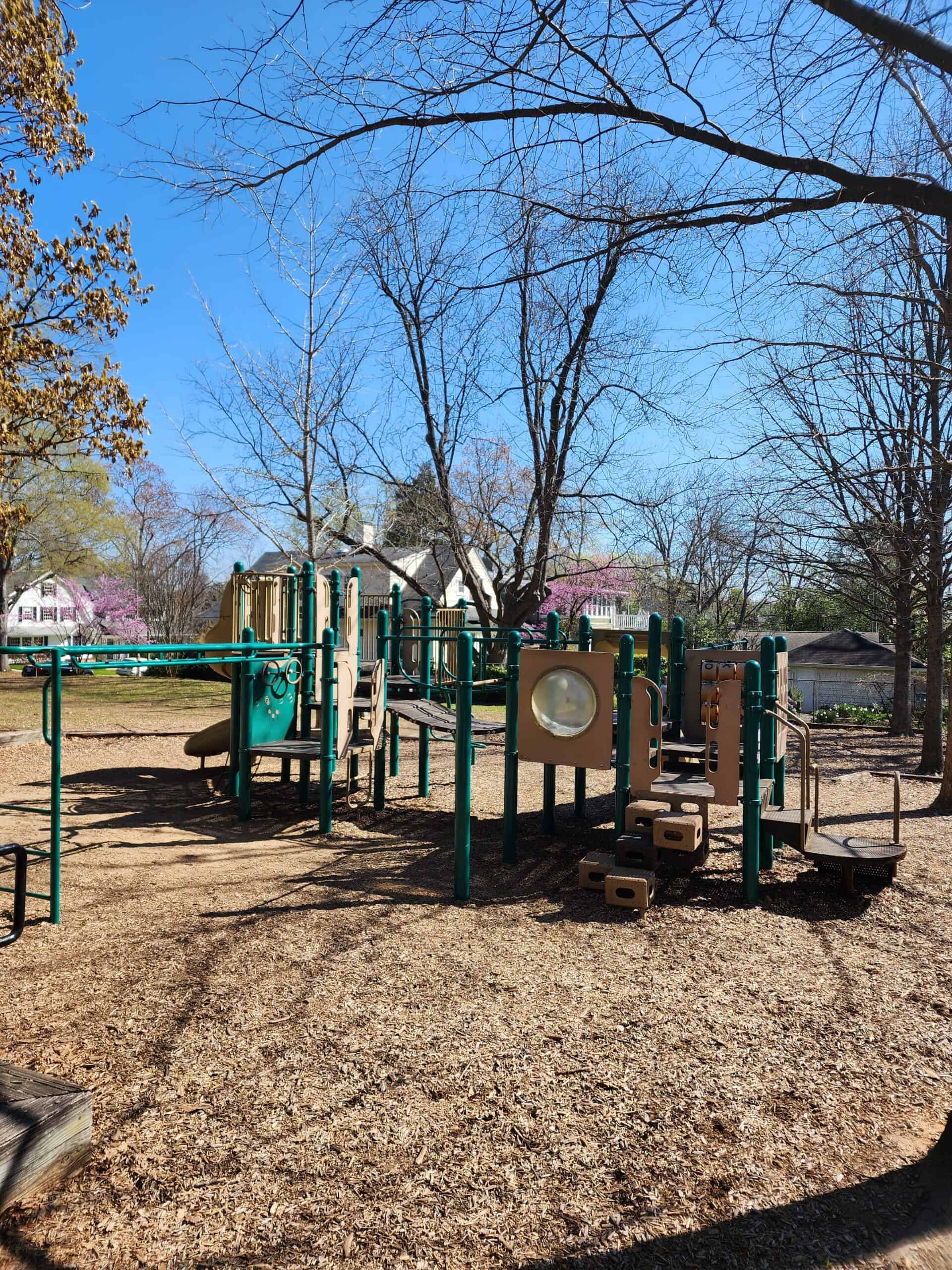 a playground with green equipment nestled among bare trees under a bright blue sky, featuring slides, climbing areas, and interactive panels, surrounded by a layer of wood chips on the ground, evoking a calm and inviting atmosphere for play.