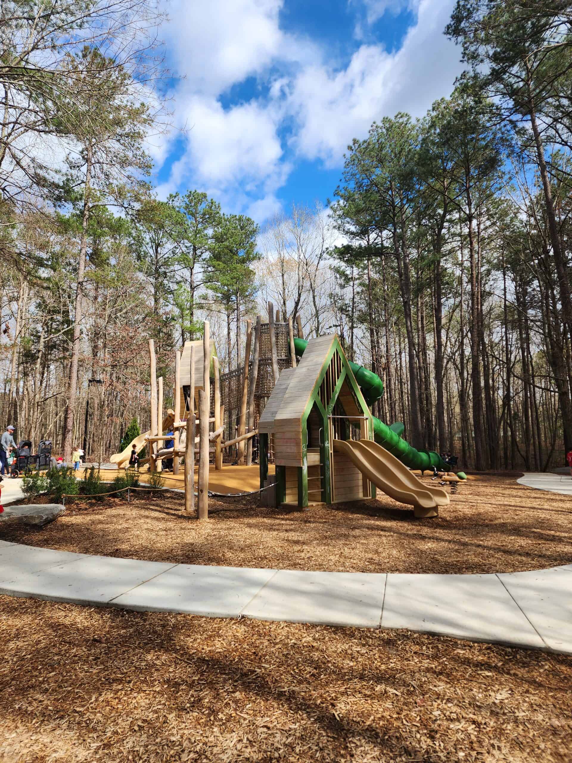 Children play on a natural wood and green plastic playground, featuring slides and climbing areas, nestled among tall pine trees with a clear blue sky overhead and a bed of wood chips underfoot