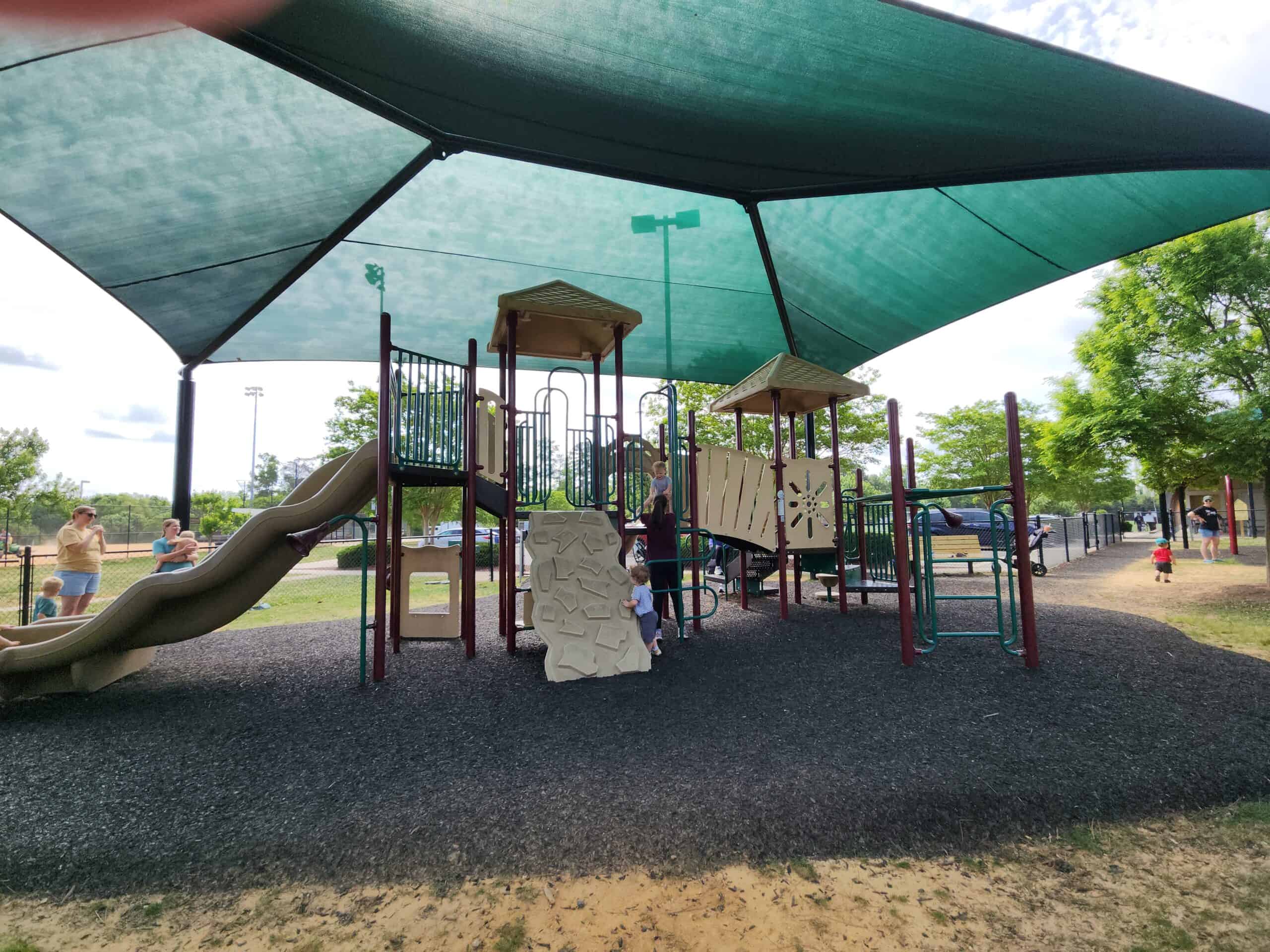 A playground with slides and climbing structures is sheltered by a large green canopy, providing ample shade. The playground surface is covered with black rubber mulch, and a few adults and children are playing and standing nearby. The surrounding area includes trees and open space, with additional people and children in the background. This image highlights a shaded playground area, ideal for staying cool during playtime.