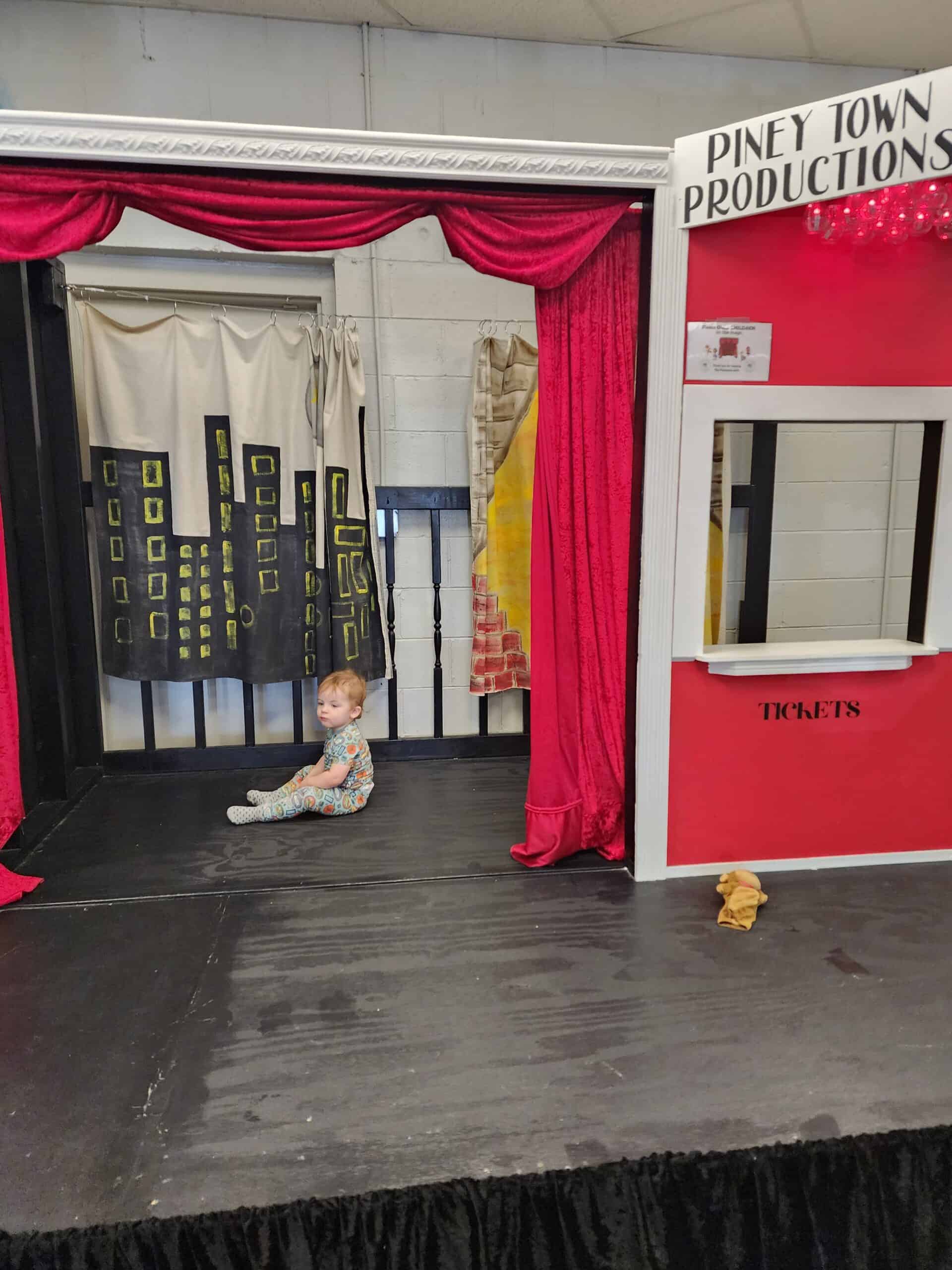 A child sits on stage under the bright lights of Piney Town Productions at the Piney Town Playhouse in Fuquay-Varina, NC, next to a ticket booth with red drapes, creating a mini theatrical experience for budding performers.