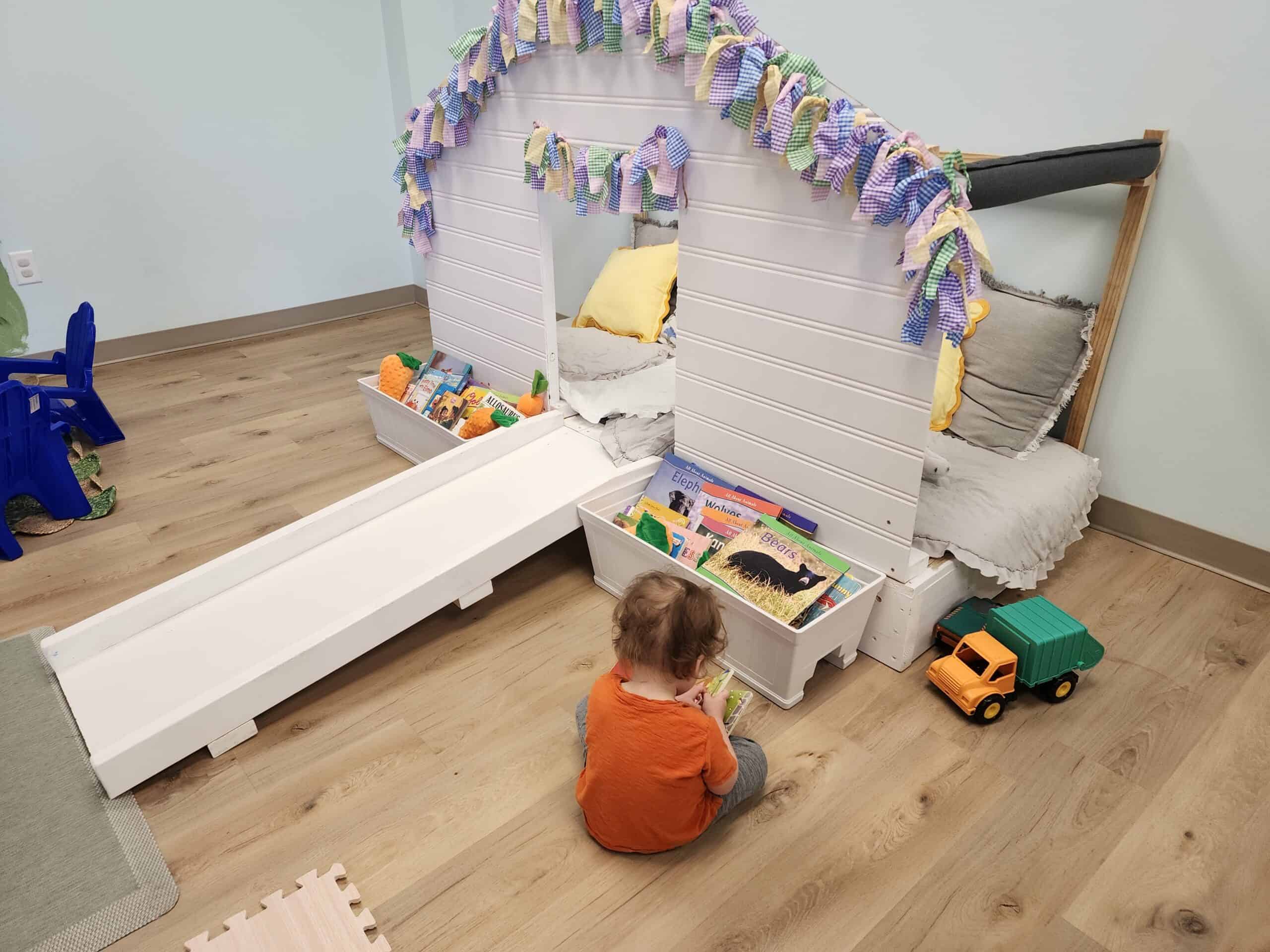 A child engrossed in reading books in the cozy book nook of Bumble Brews indoor playground in Raleigh, NC, with a soft fabric garland overhead and a gentle slide, encouraging quiet time and reading in a playful setting.