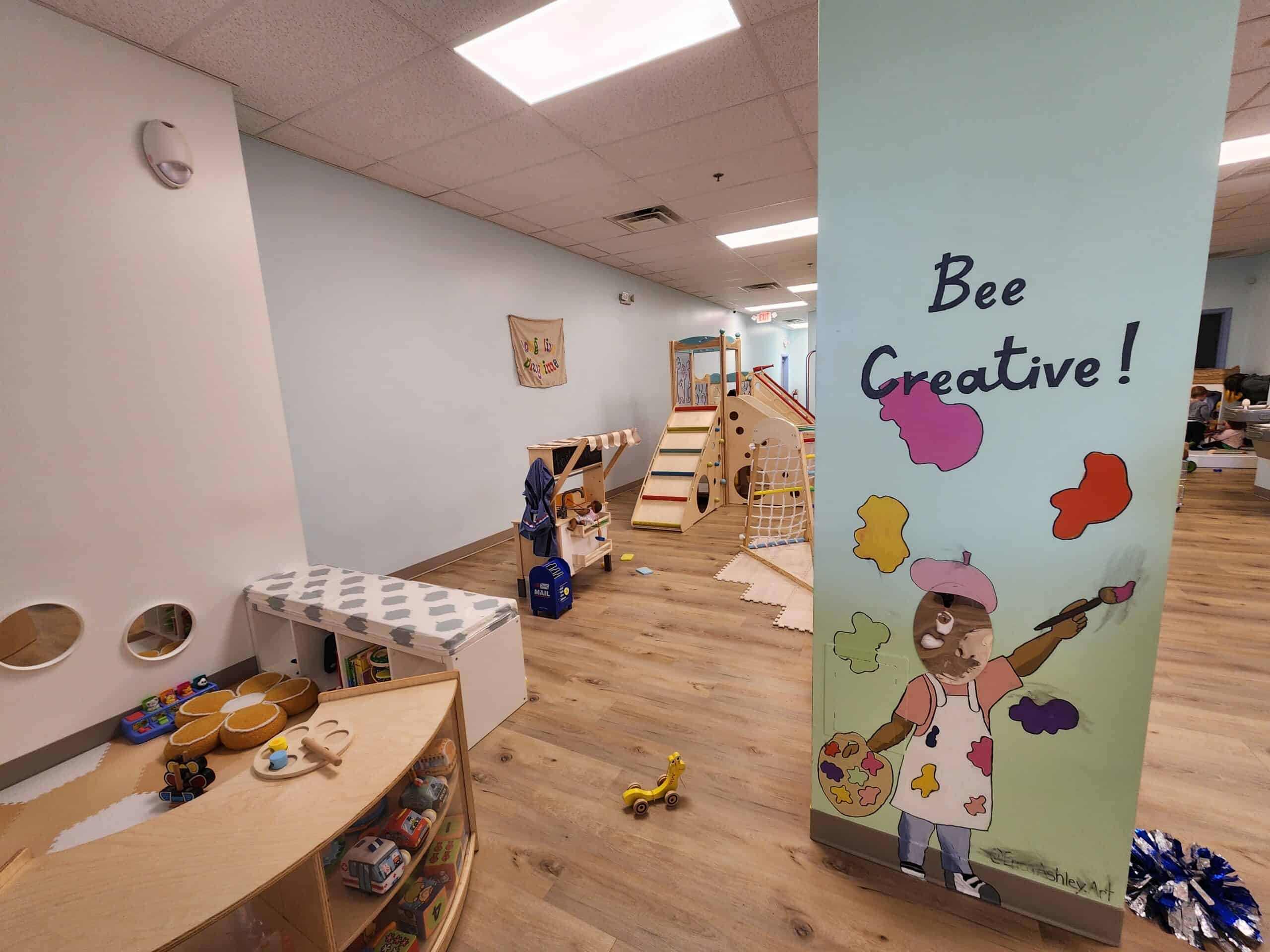 Creative play area at Bumble Brews indoor playground in Raleigh, NC, featuring a playful mural with 'Bee Creative!' slogan, and a variety of interactive toys and structures for children's imaginative play.