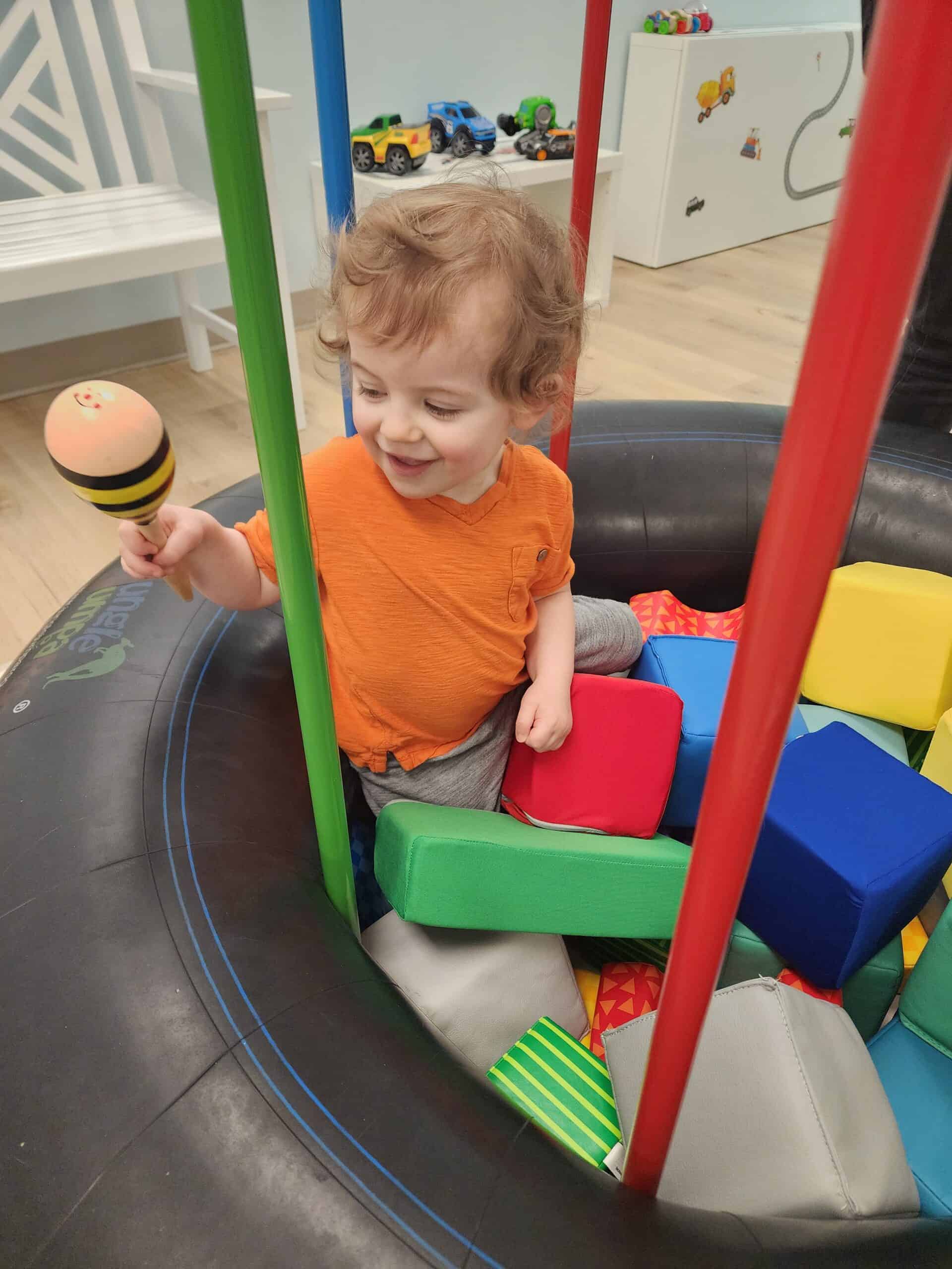 A joyful toddler with curly hair holding a toy maraca inside a colorful padded play area at Bumble Brews indoor playground in Raleigh, NC, surrounded by soft blocks and vibrant play structures.